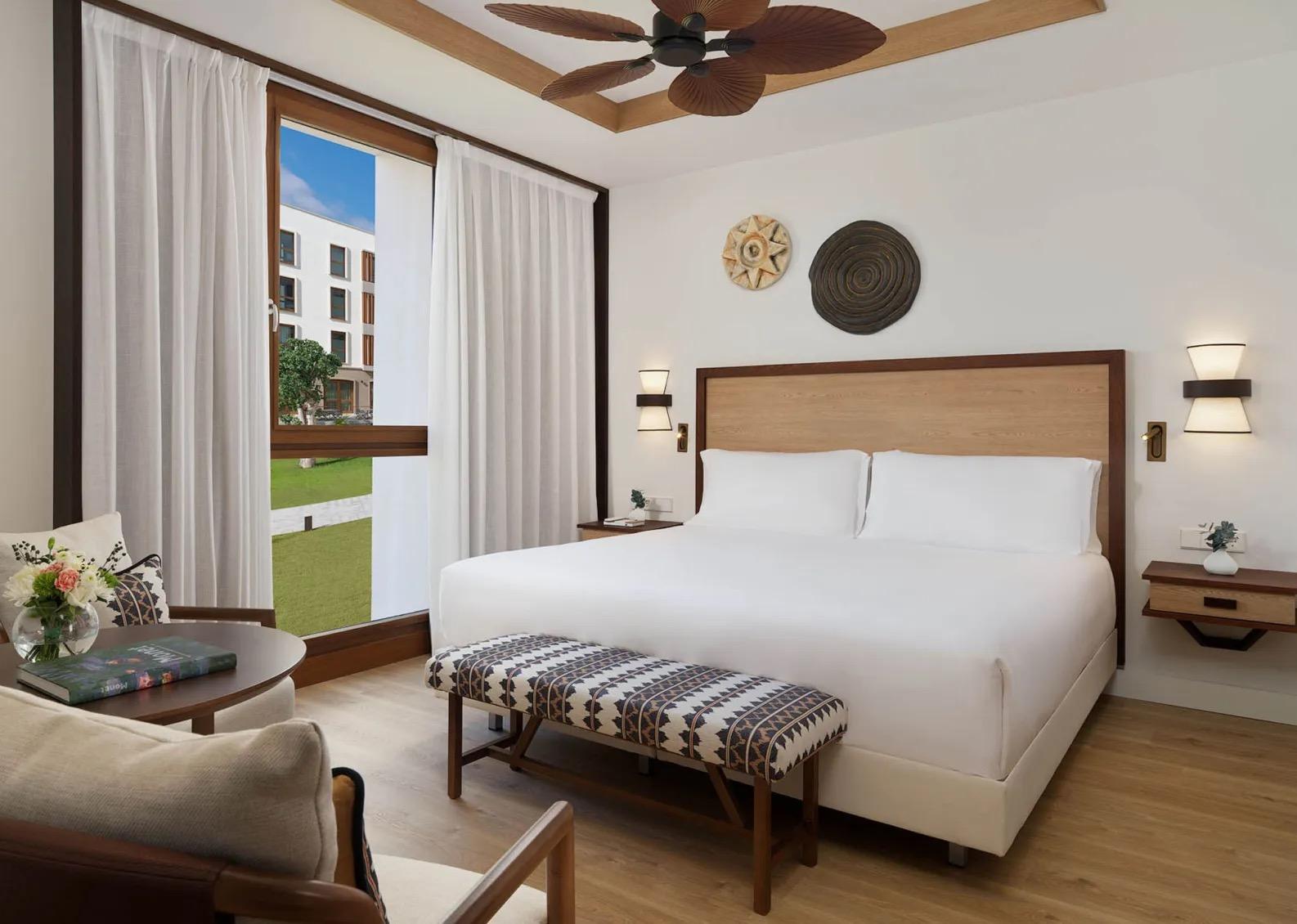 A hotel bedroom with a wood-trimmed bed beneath a rattan ceiling fan, curtains open to a window overlooking a lawn and a modern building.