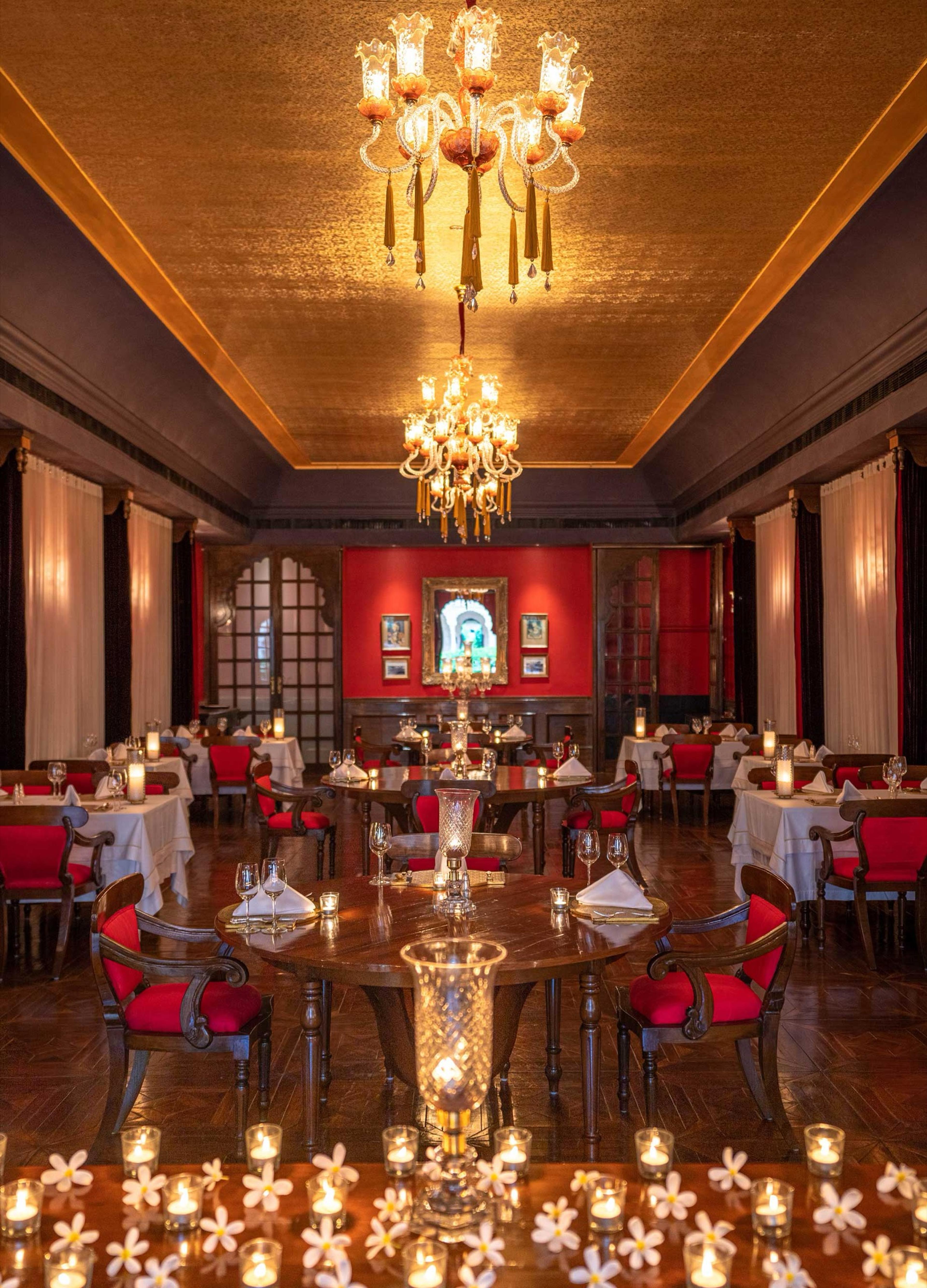 a red dining hall with two crystal chandeliers, rows of tables laid with glassware, and candles and frangipani flowers in the foreground