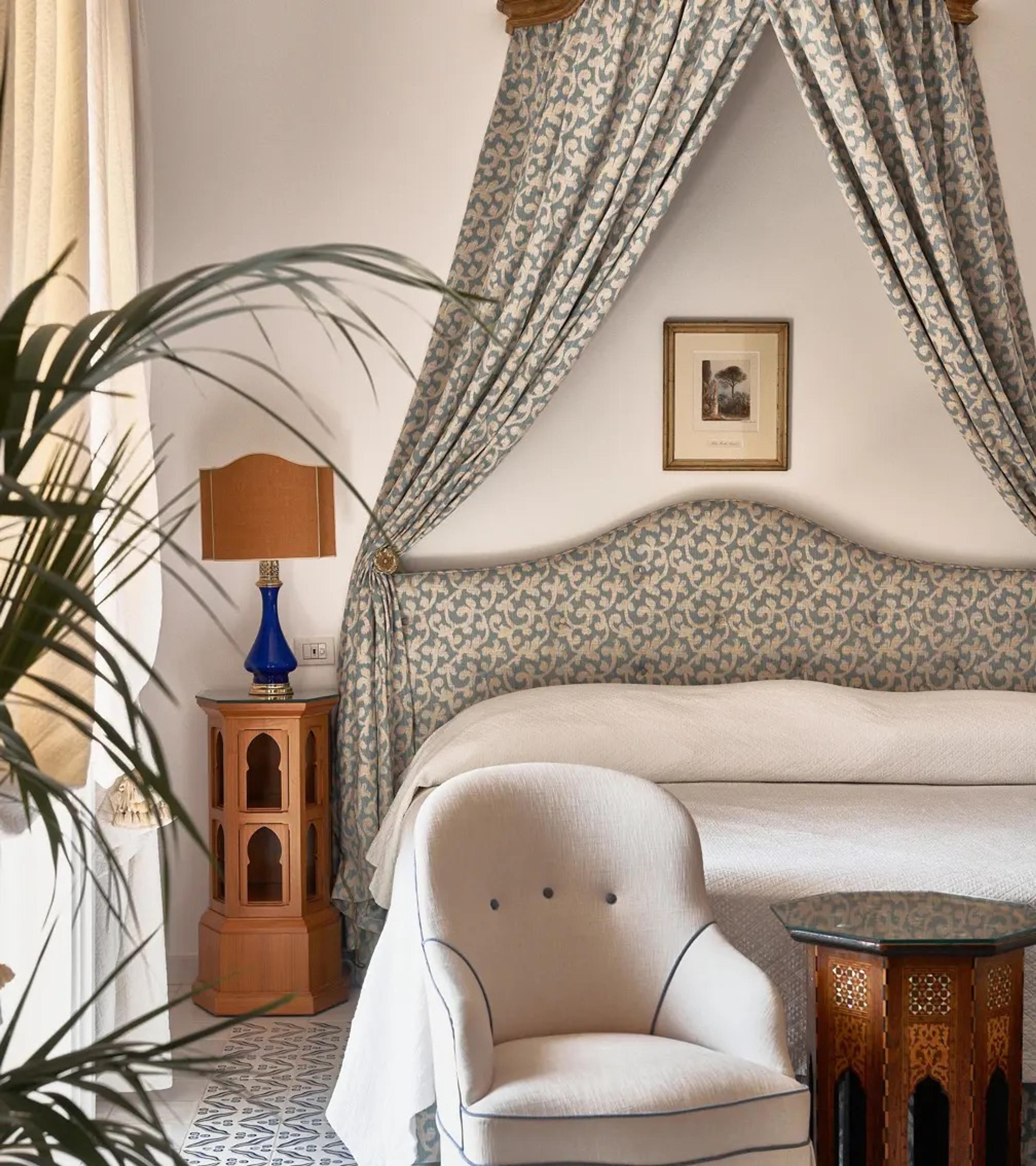 A bed with a woven headboard under a draped canopy, a lamp and a picture on the wall beside it.