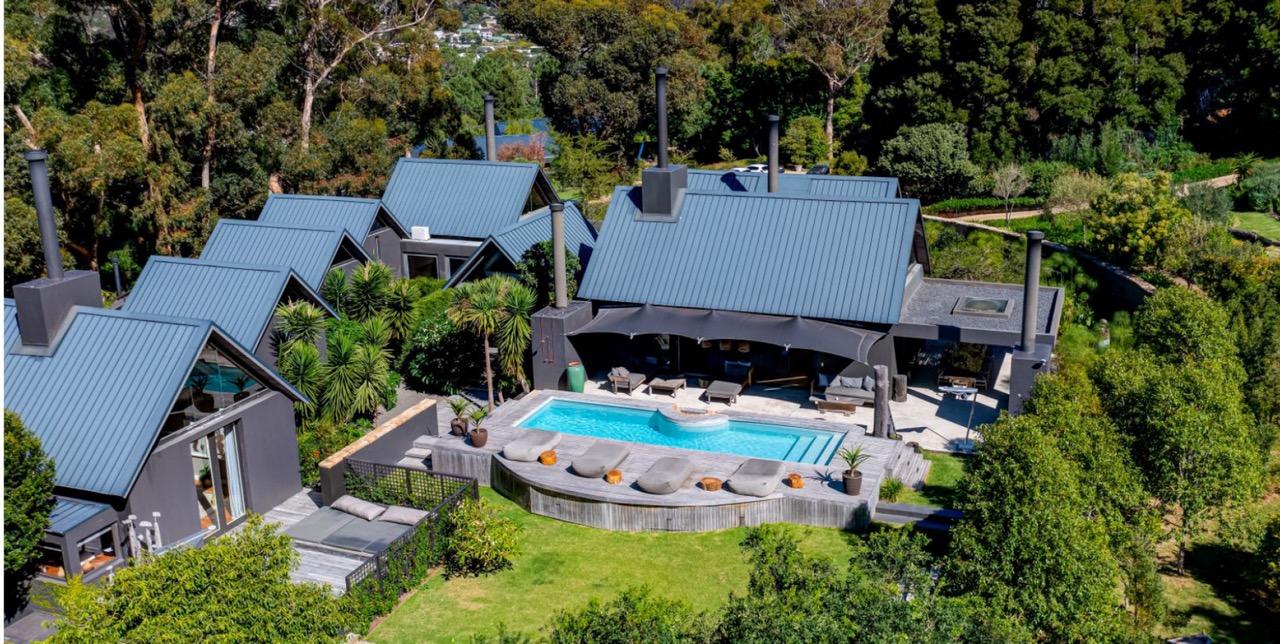 An aerial view of a cluster of dark-roofed buildings around a swimming pool with a shaded lounge area, surrounded by trees and lawn.