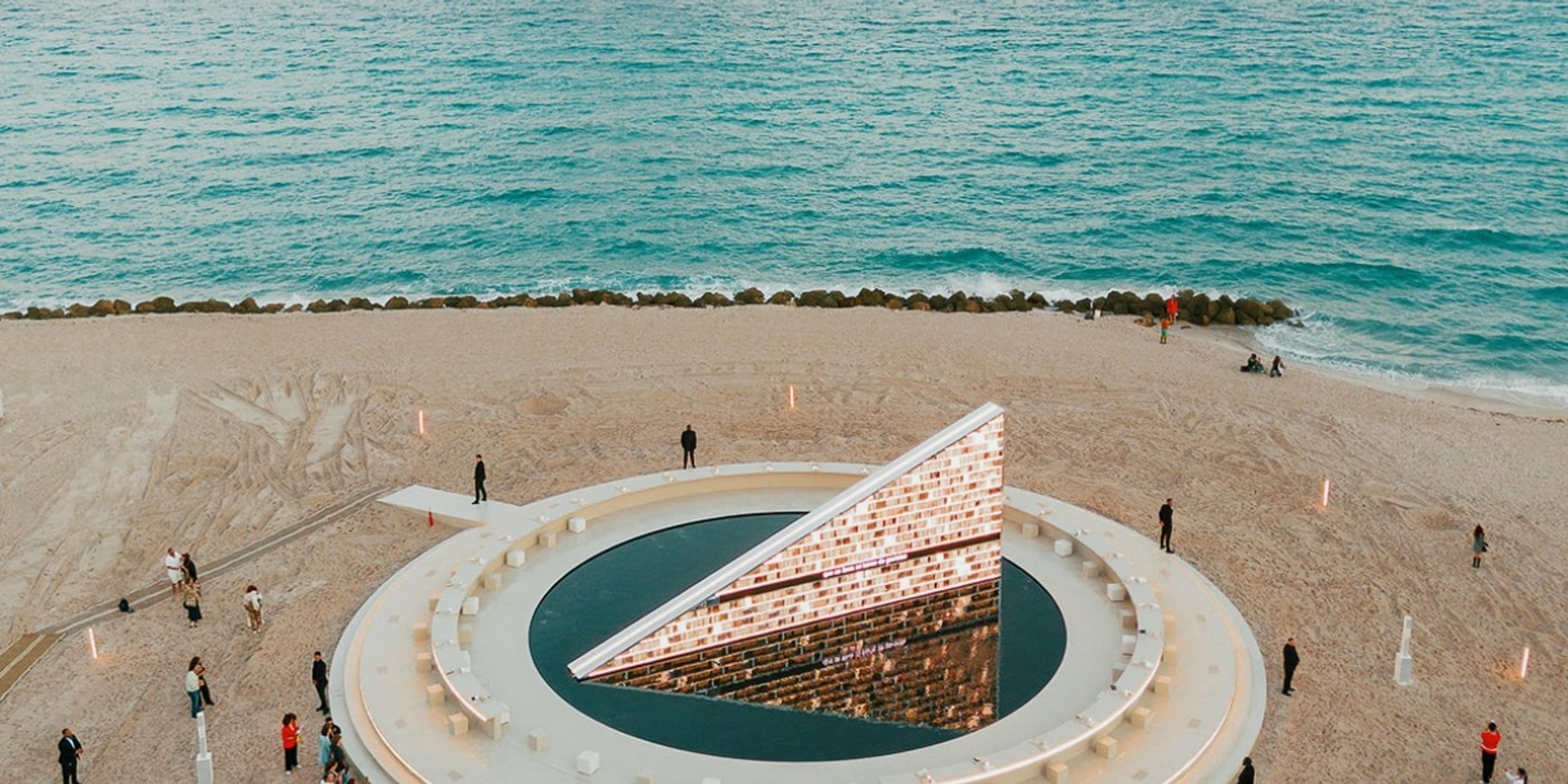 An aerial view of a round pool on the sand with a tilted, lit, brick-patterned triangular monument standing in it, people walking around the edge and the sea beyond.