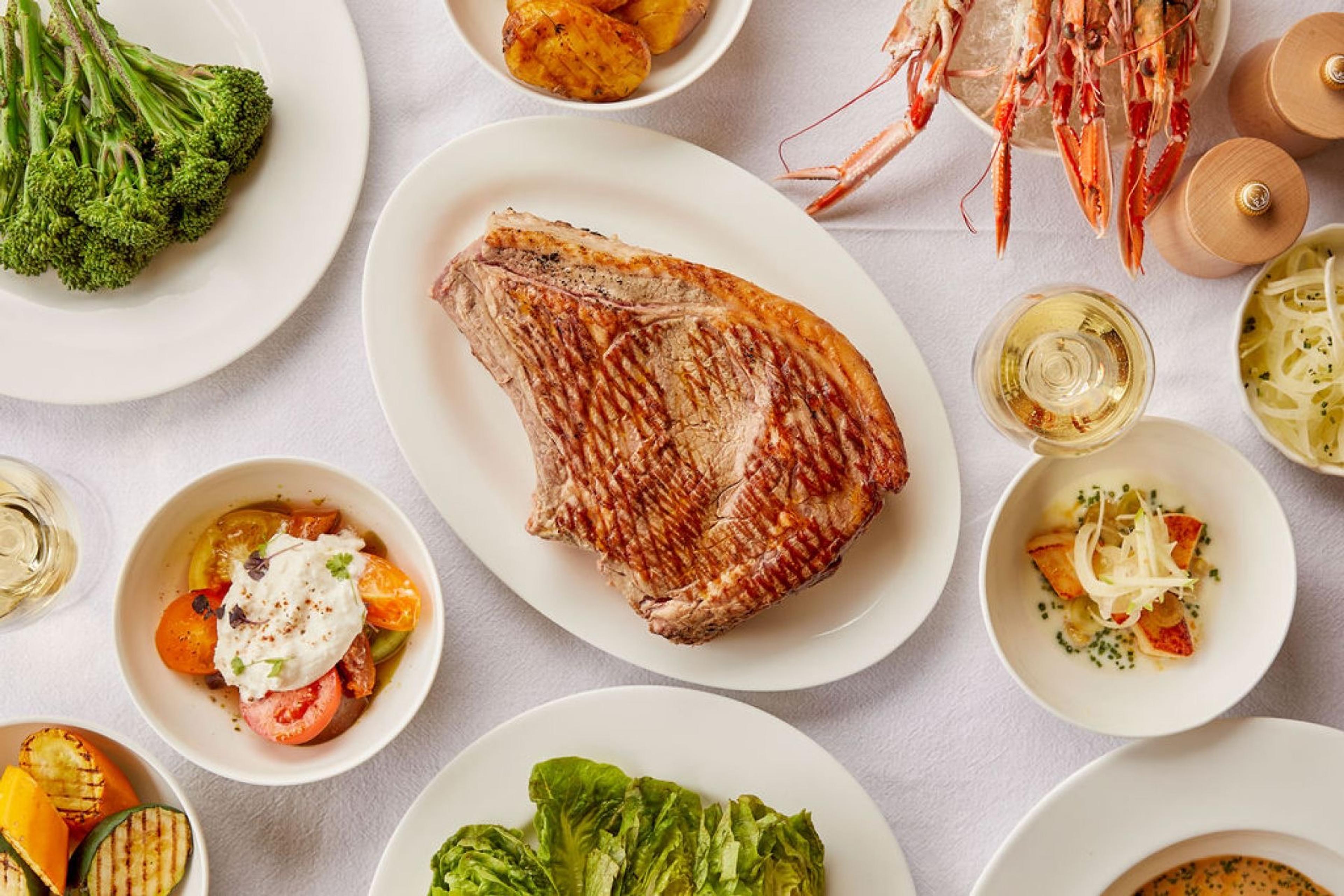 An overhead view of a shared table with grilled pork chop, langoustines, broccoli, burrata and tomatoes, and bowls of sauce and salad.