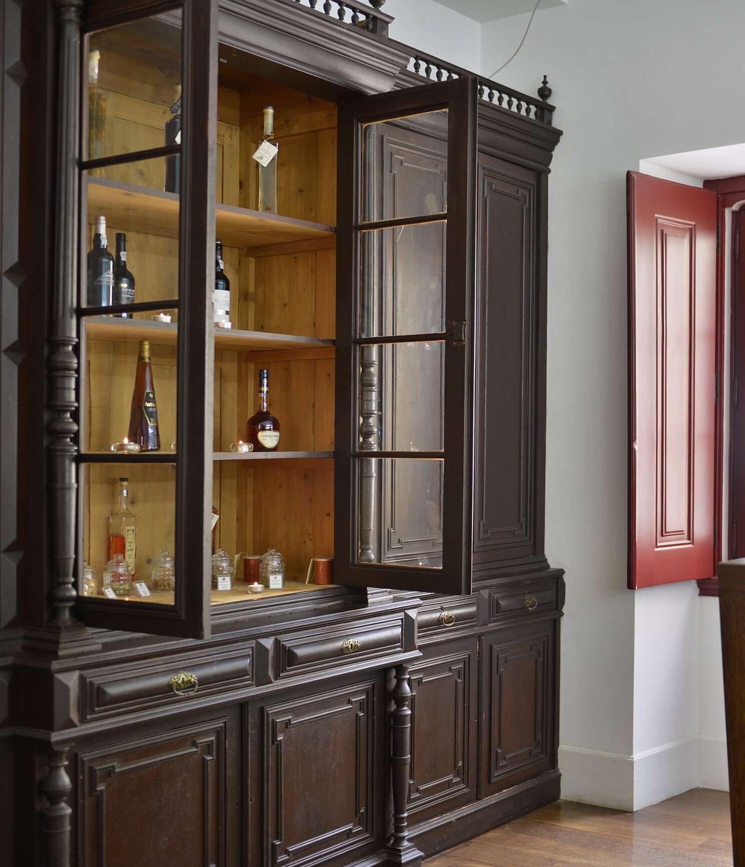 A dark wooden cabinet with glass doors, one open, showing bottles and jars on pine shelves.