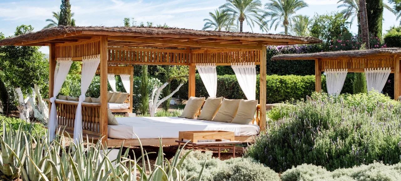 A single thatched cabana with white curtains tied back and cushions along its bench, set among aloe and lavender.