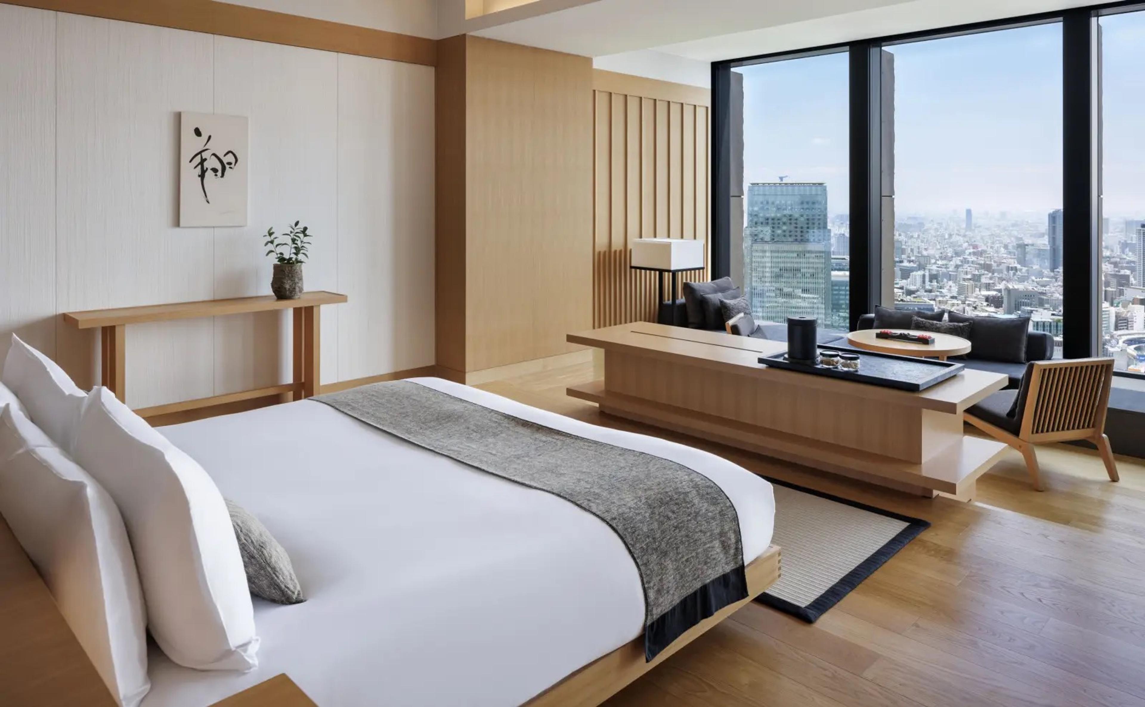 A bedroom with a white-made bed, a wooden console table, and a built-in bench seat facing floor-to-ceiling windows over the city.