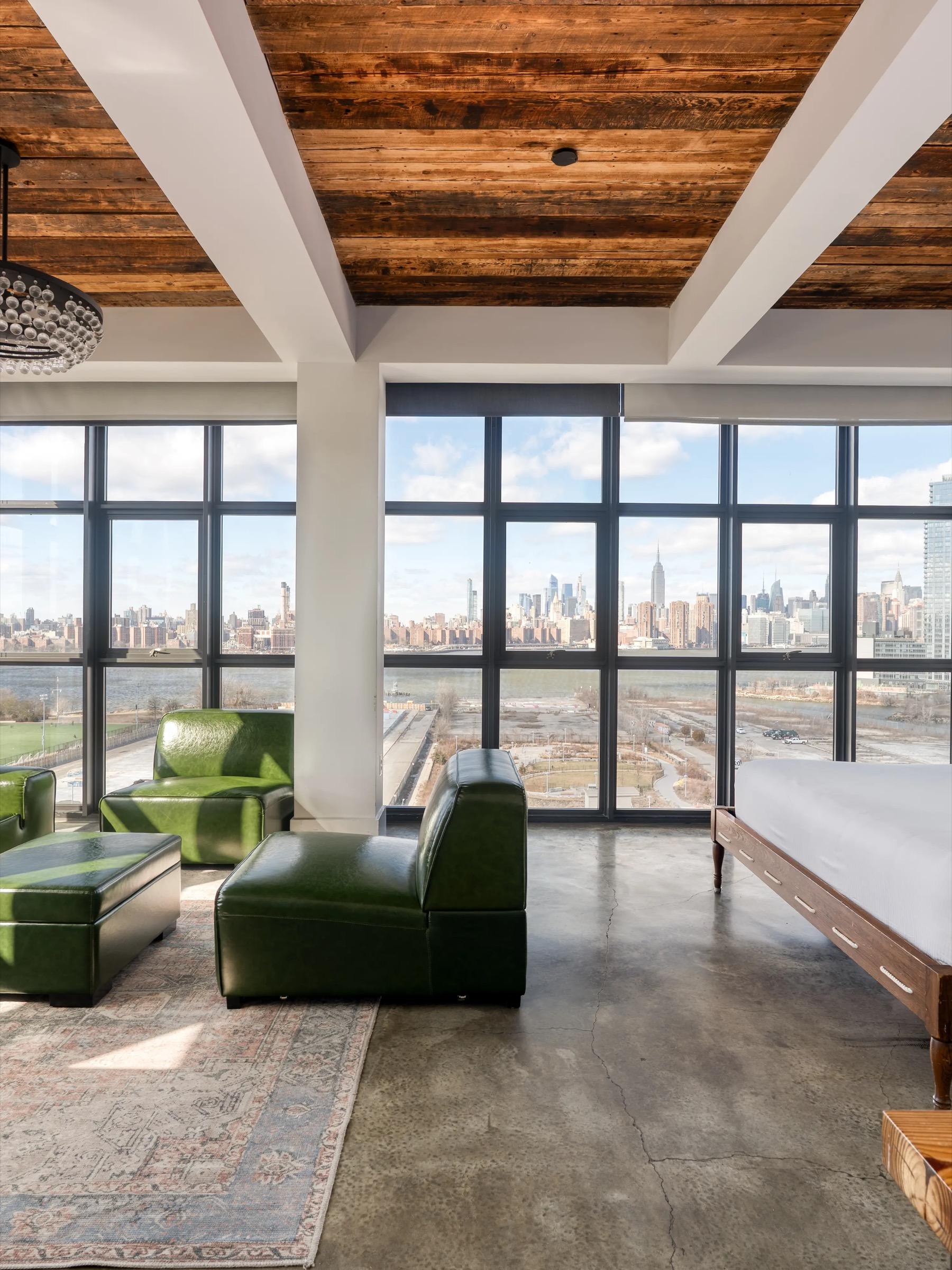 A double height hotel loft with a wood beamed ceiling, green leather lounge chairs on a patterned rug, and a wall of windows overlooking the Brooklyn waterfront and the Manhattan skyline.