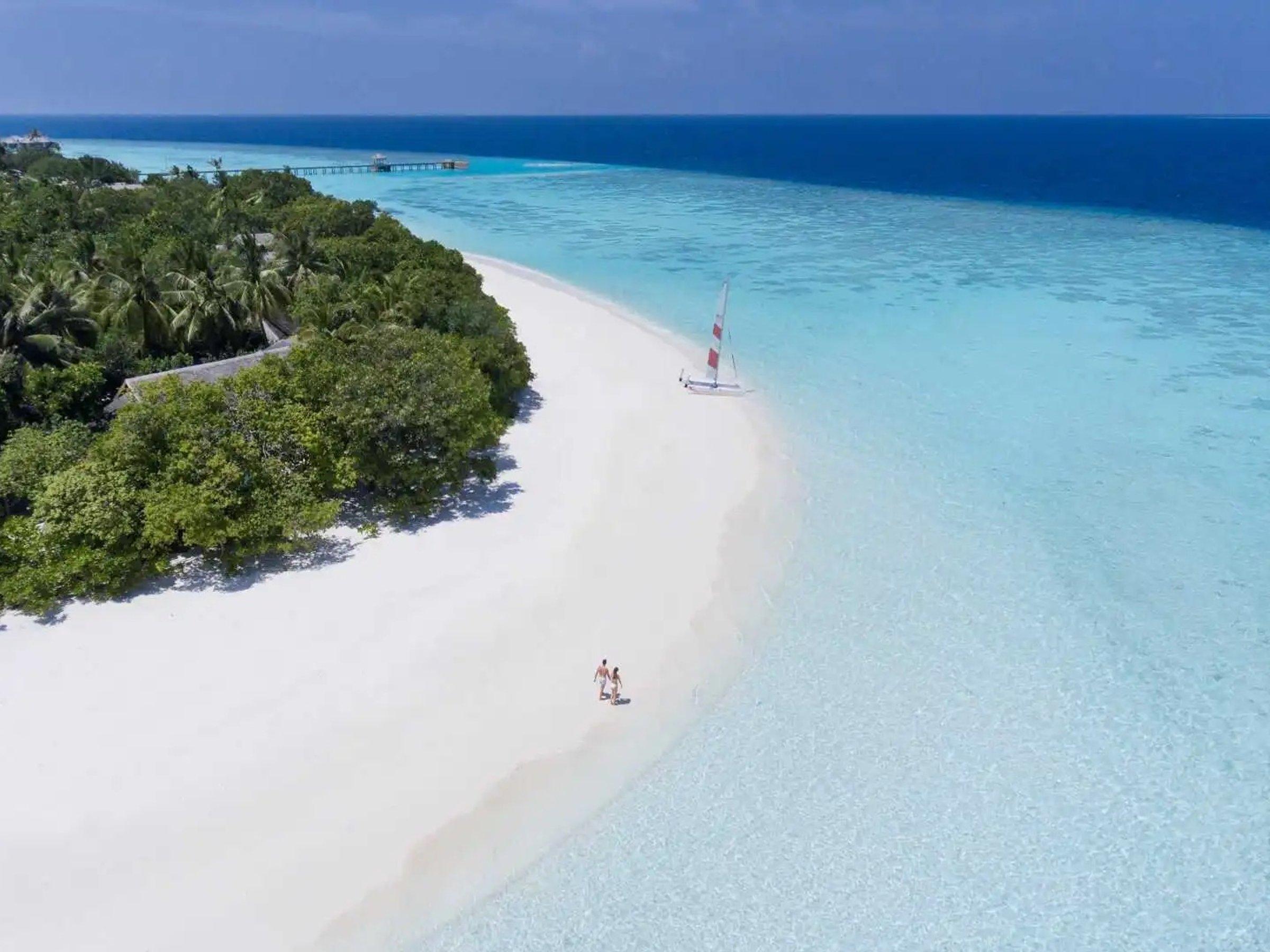 A curve of white sand on a Baa Atoll island, two people walking at the water's edge and a small catamaran pulled up on the beach