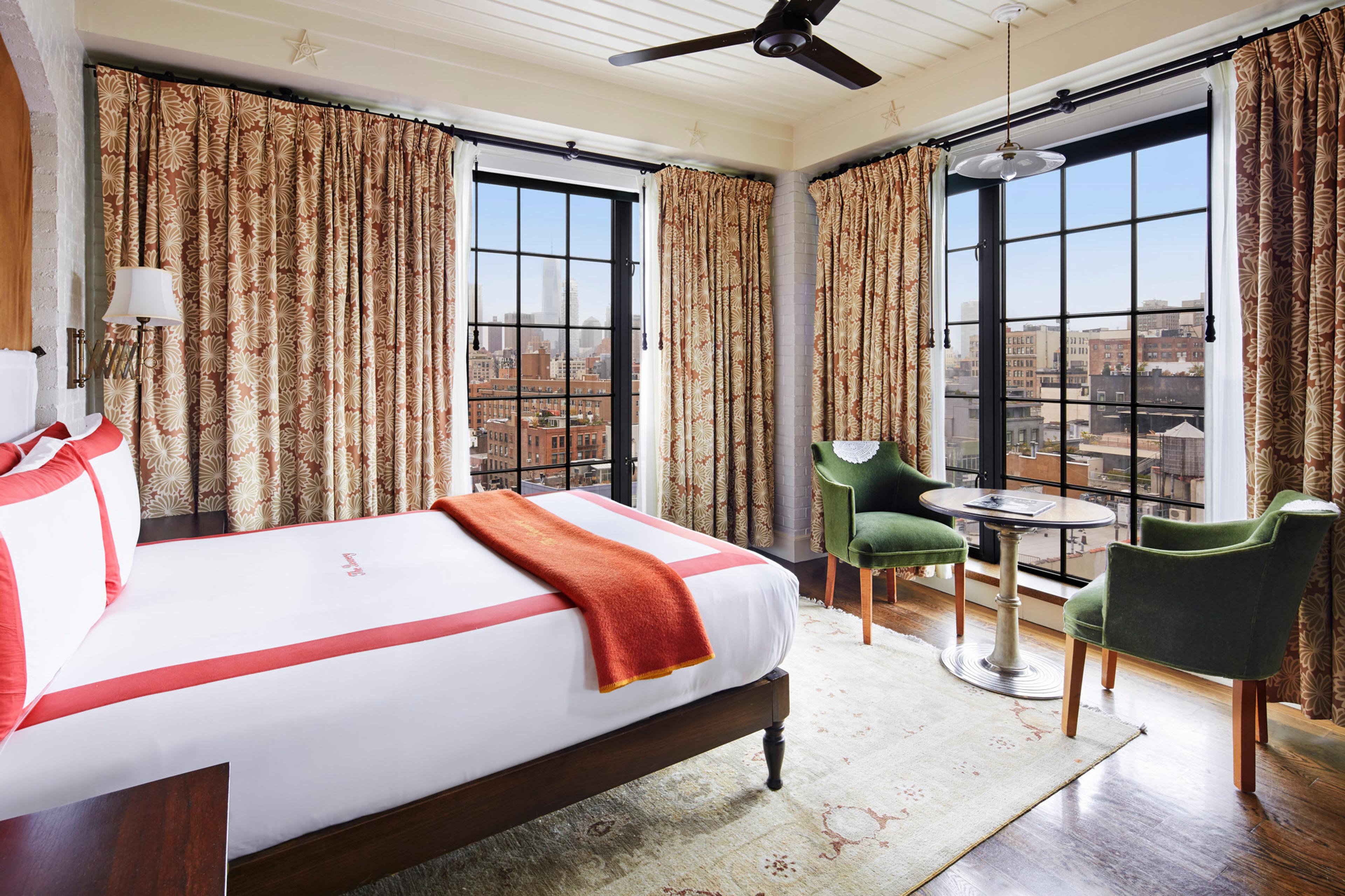 A hotel room with a queen bed, a round table and two chairs by large windows looking out over the city.