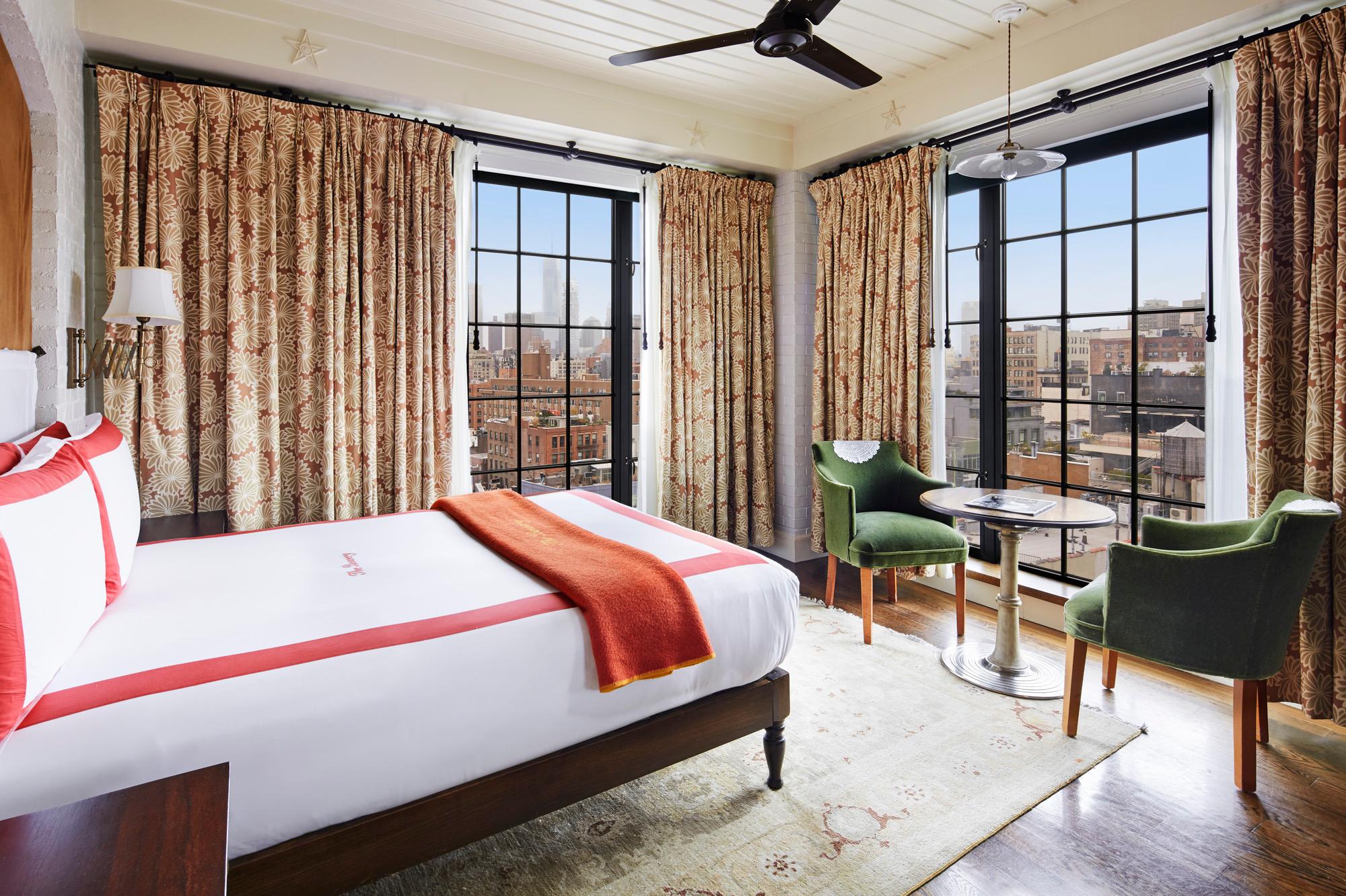 A hotel room with a queen bed, a round table and two chairs by large windows looking out over the city.