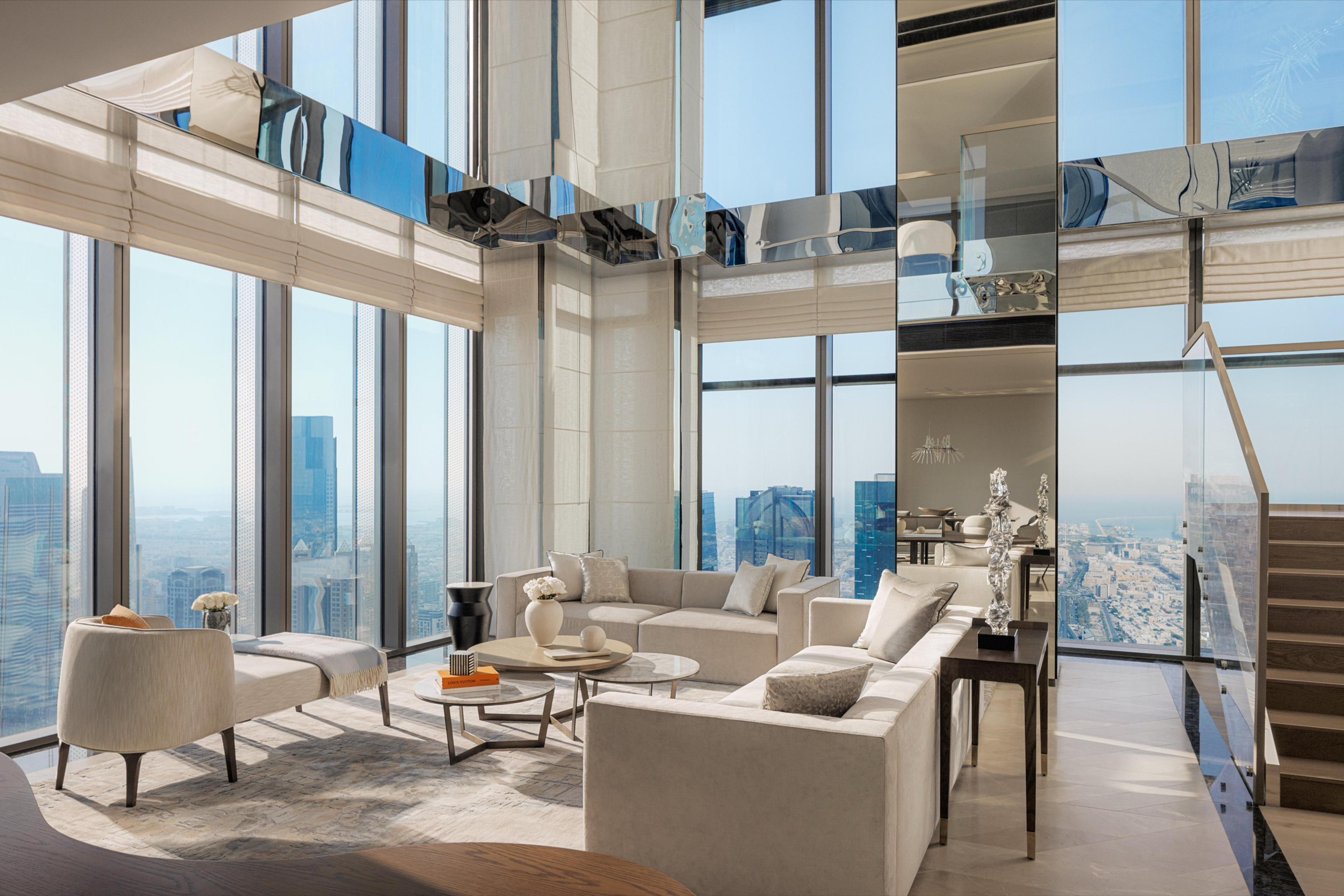 A double-height living room with floor-to-ceiling windows on two sides, pale sofas and a bench, a spiral staircase, and a wide view over a daytime city skyline.