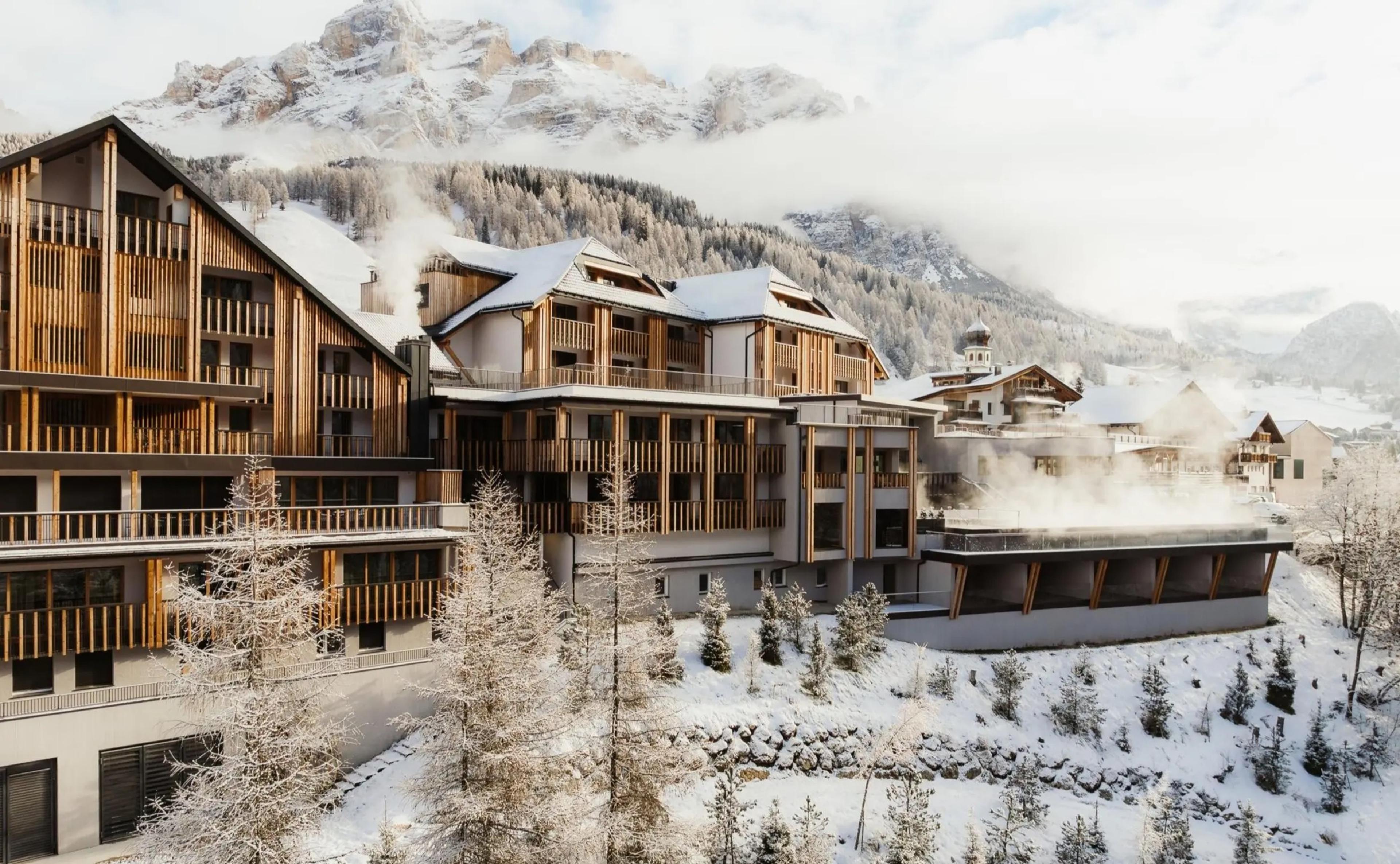 a large timber-clad hotel building with snow covered balconies, a steaming spa pavilion beside it, and a sharp mountain peak rising behind