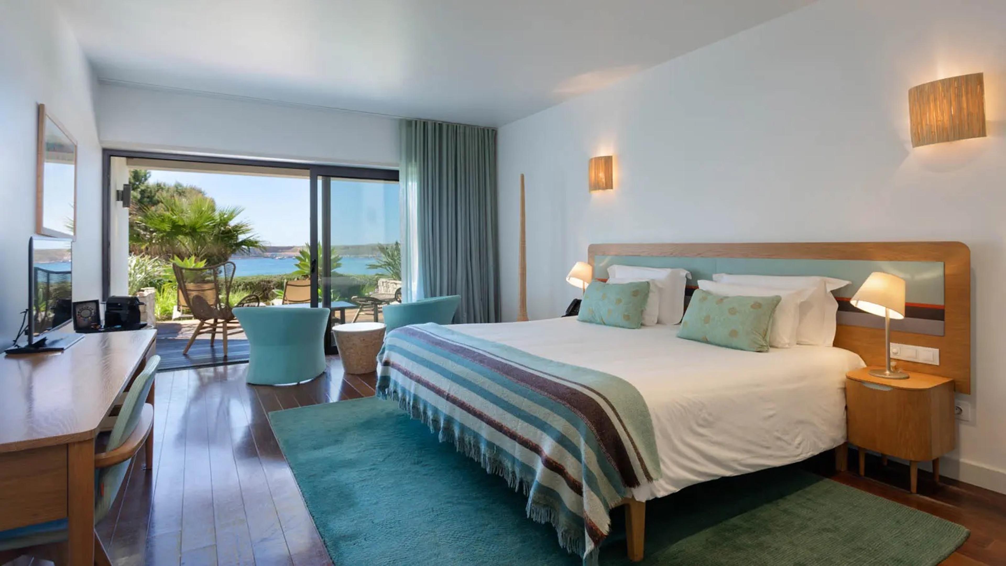 A double bedroom with a teal headboard and striped throw, with sliding glass doors onto a terrace and the sea beyond.