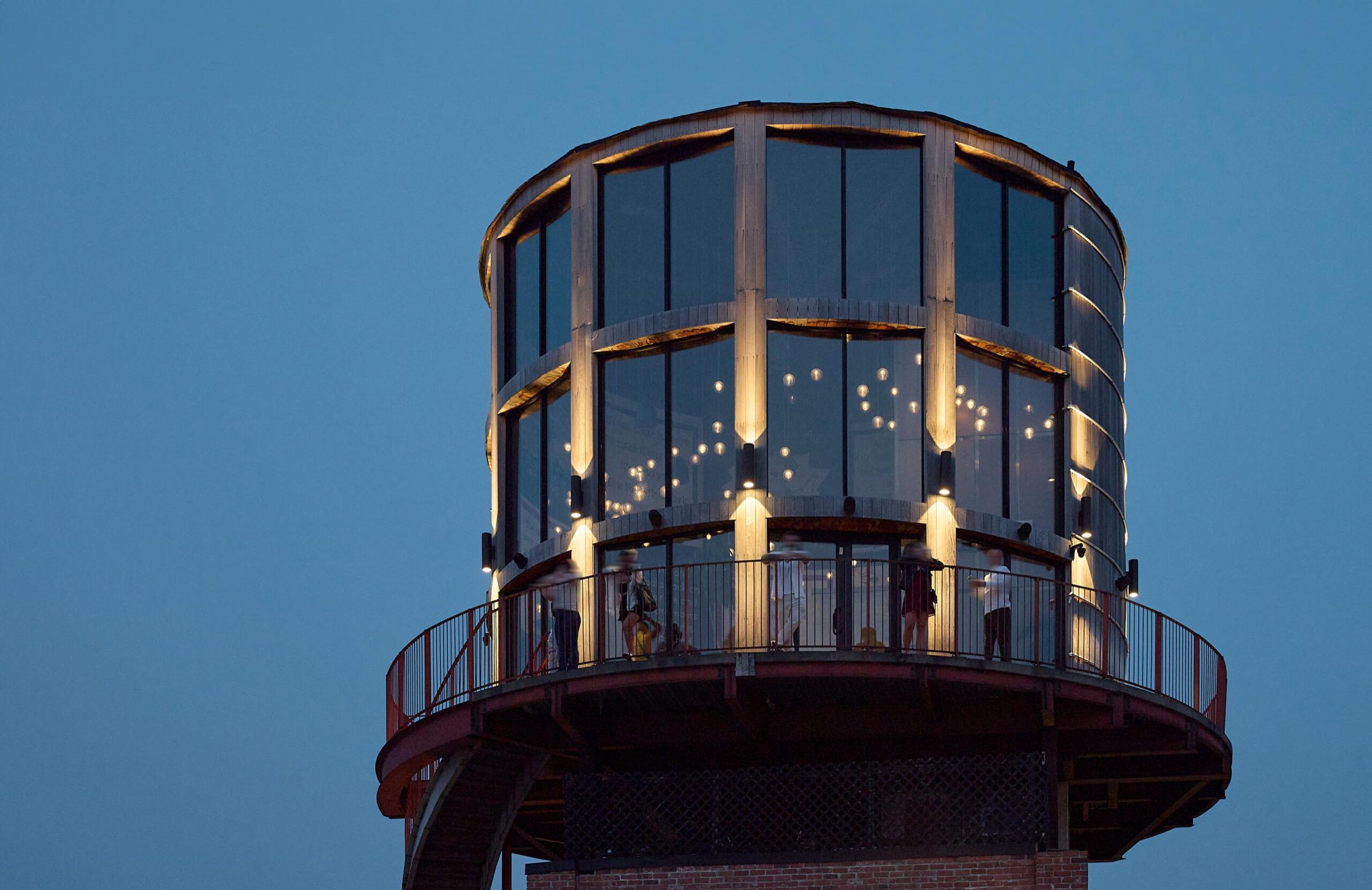 A glass-walled water tower structure lit from within at dusk, with a handful of people visible on its wraparound deck against a darkening blue sky.