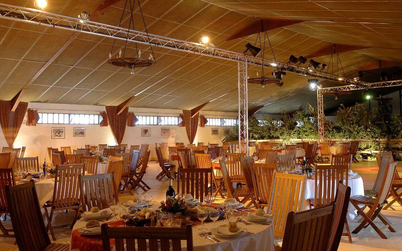 Long wooden tables laid with white cloths, glassware and bread rolls inside a barn-style dining room strung with lights.