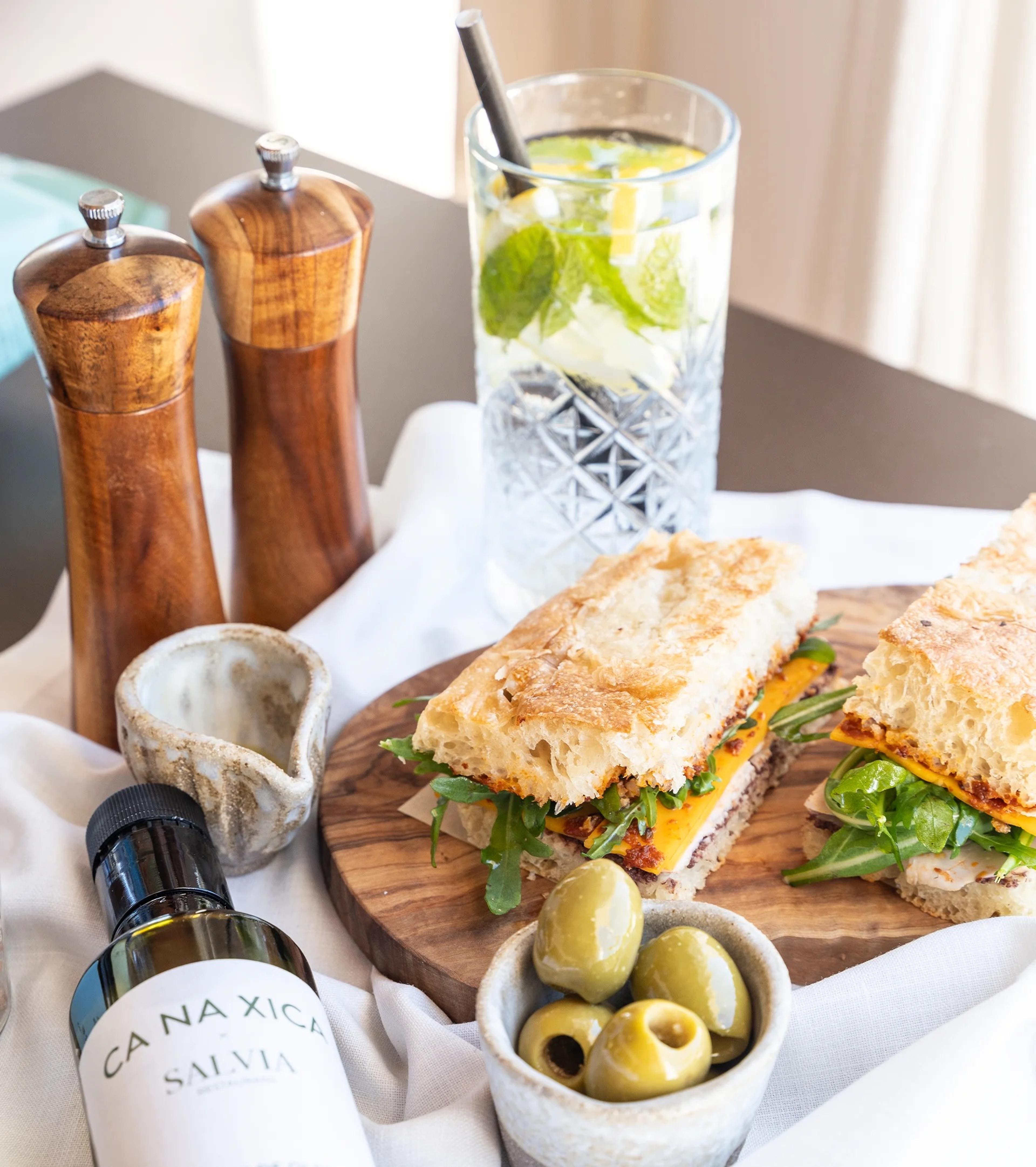 Two halves of a filled baguette sandwich on a wooden board, next to a bowl of olives, a glass of iced mint tea, pepper grinders, and a bottle of Ca Na Xica olive oil.