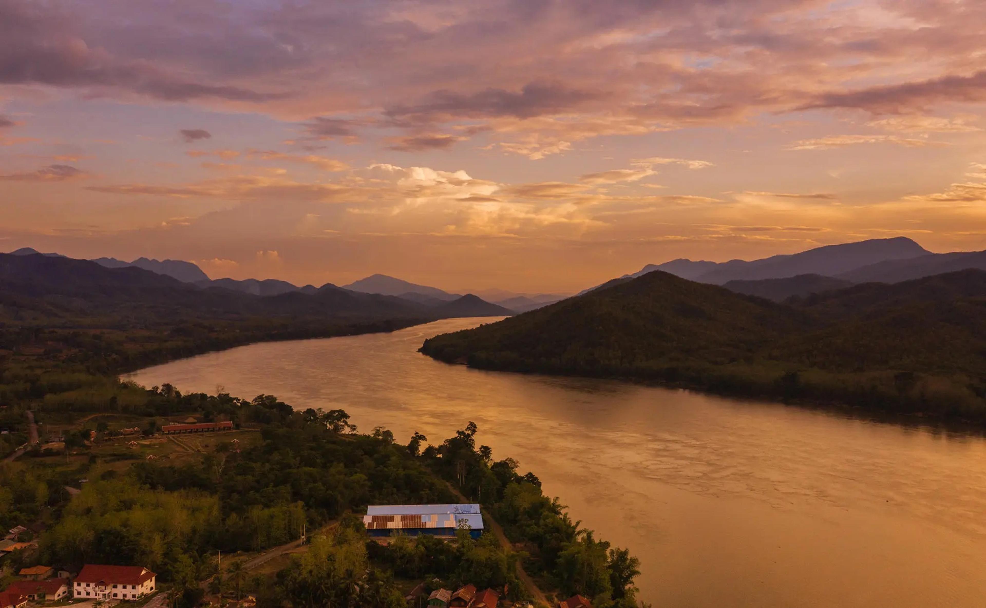 An aerial view at sunset of the Mekong river curving between forested hills, with rooftops clustered at the water's edge.