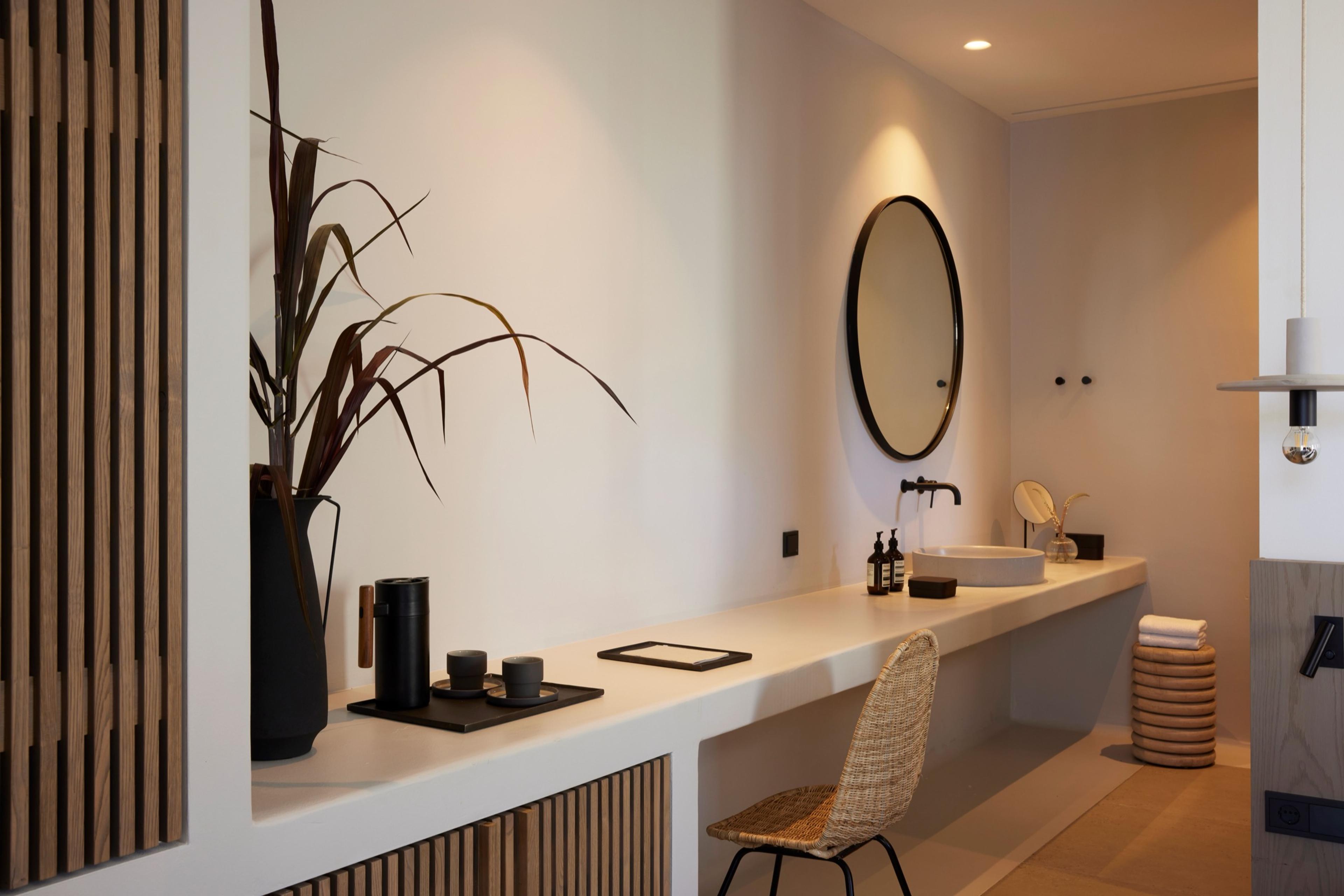A bathroom vanity with a round mirror, a sink and a woven chair, beside a shelf with a plant.