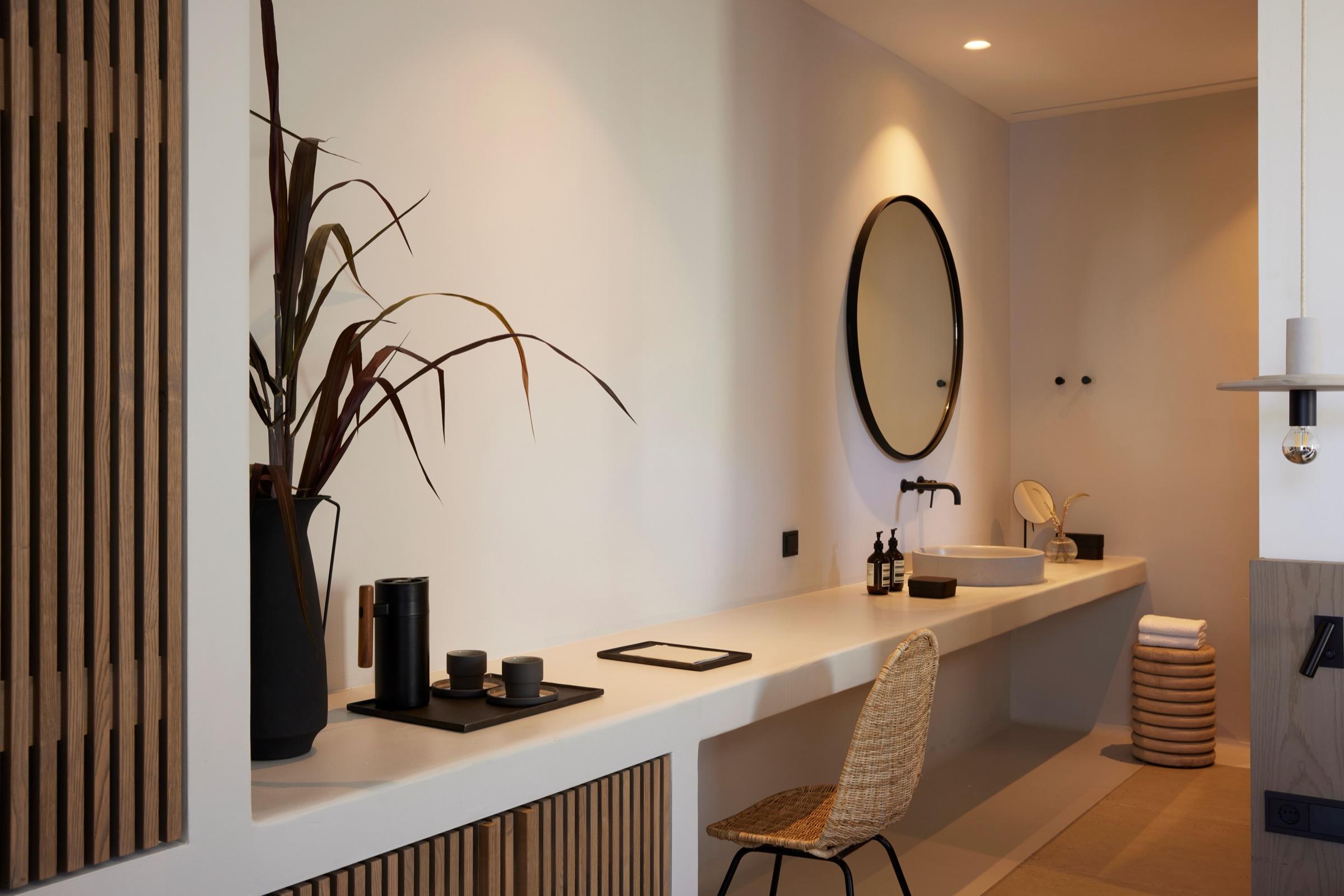 A bathroom vanity with a round mirror, a sink and a woven chair, beside a shelf with a plant.