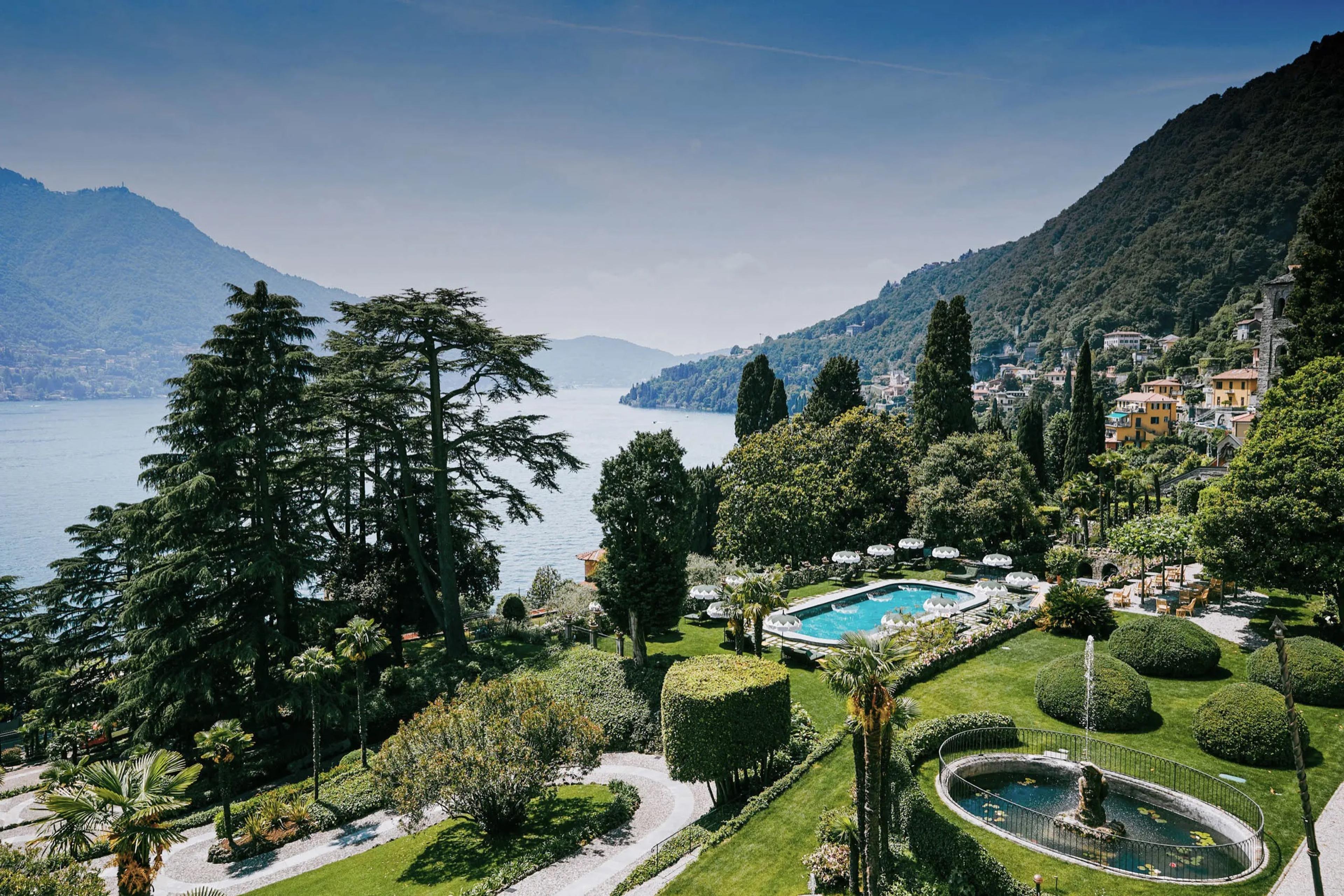 An aerial view of terraced lawns, a pool and a fountain above a lake, with a lakeside town and mountains in the distance.