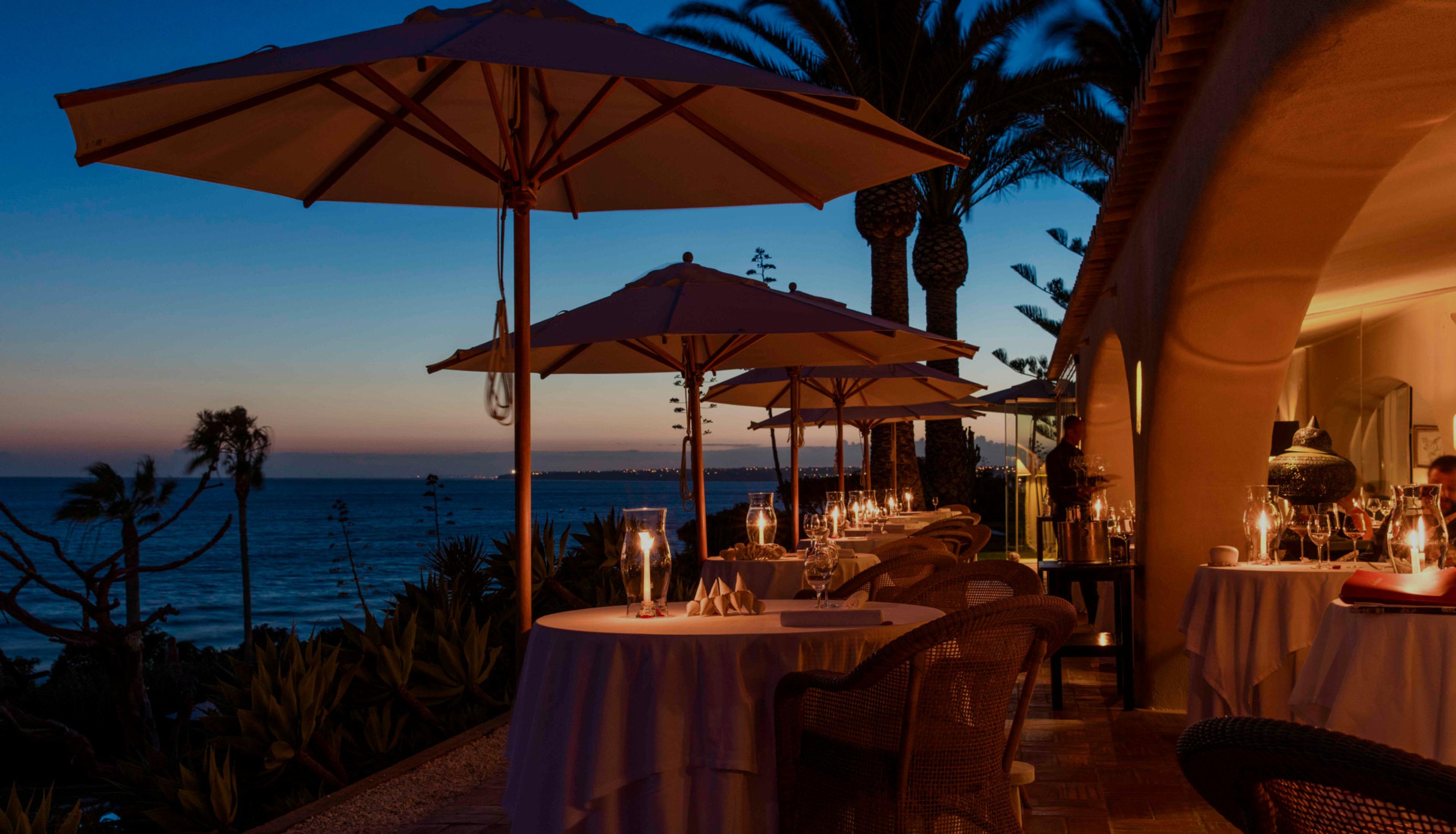 Tables under parasols on Vila Joya's terrace at dusk, the sea beyond