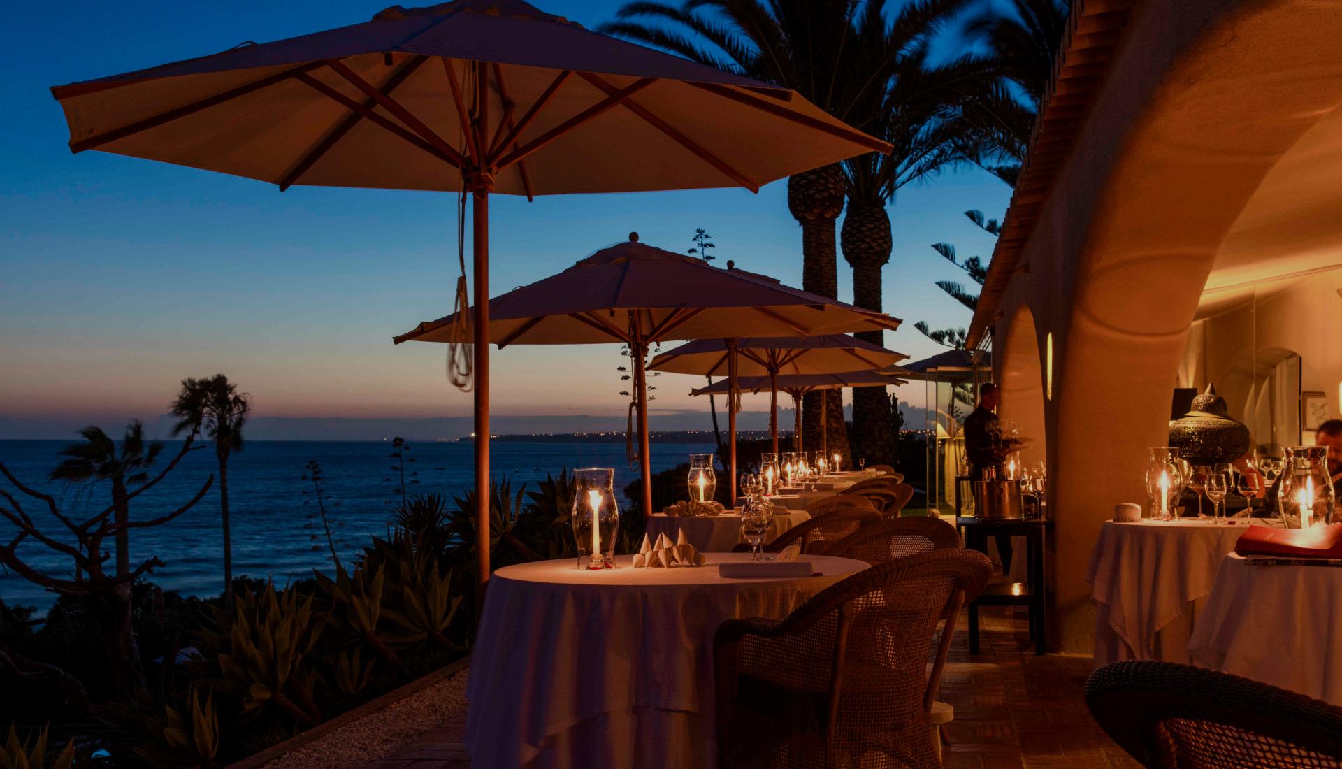 Tables under parasols on Vila Joya's terrace at dusk, the sea beyond