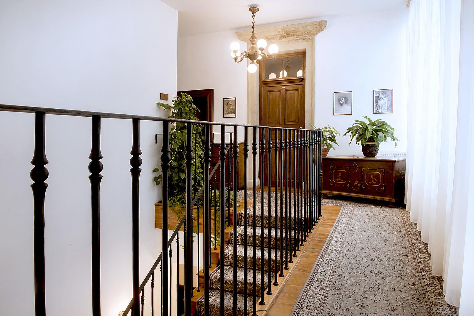 A staircase landing with a wrought-iron railing, potted plants and framed photographs on a white wall.