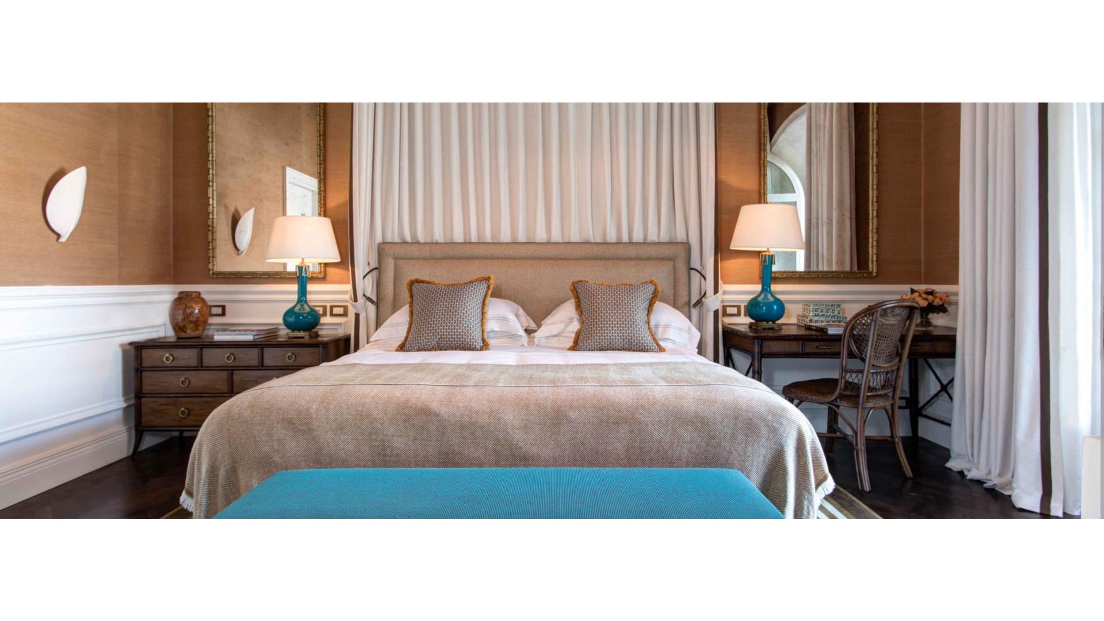 A bedroom with a canopied bed in taupe linen, flanked by two blue ceramic lamps on dark wood tables, with sheer curtains and a teal bench at the foot of the bed.