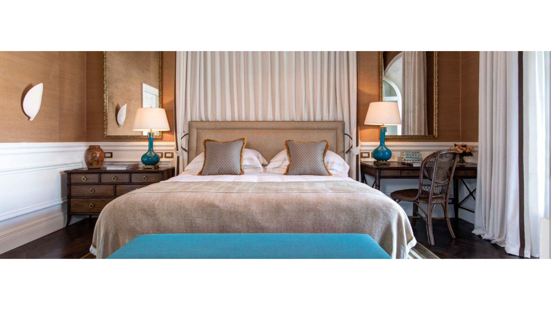 A bedroom with a canopied bed in taupe linen, flanked by two blue ceramic lamps on dark wood tables, with sheer curtains and a teal bench at the foot of the bed.