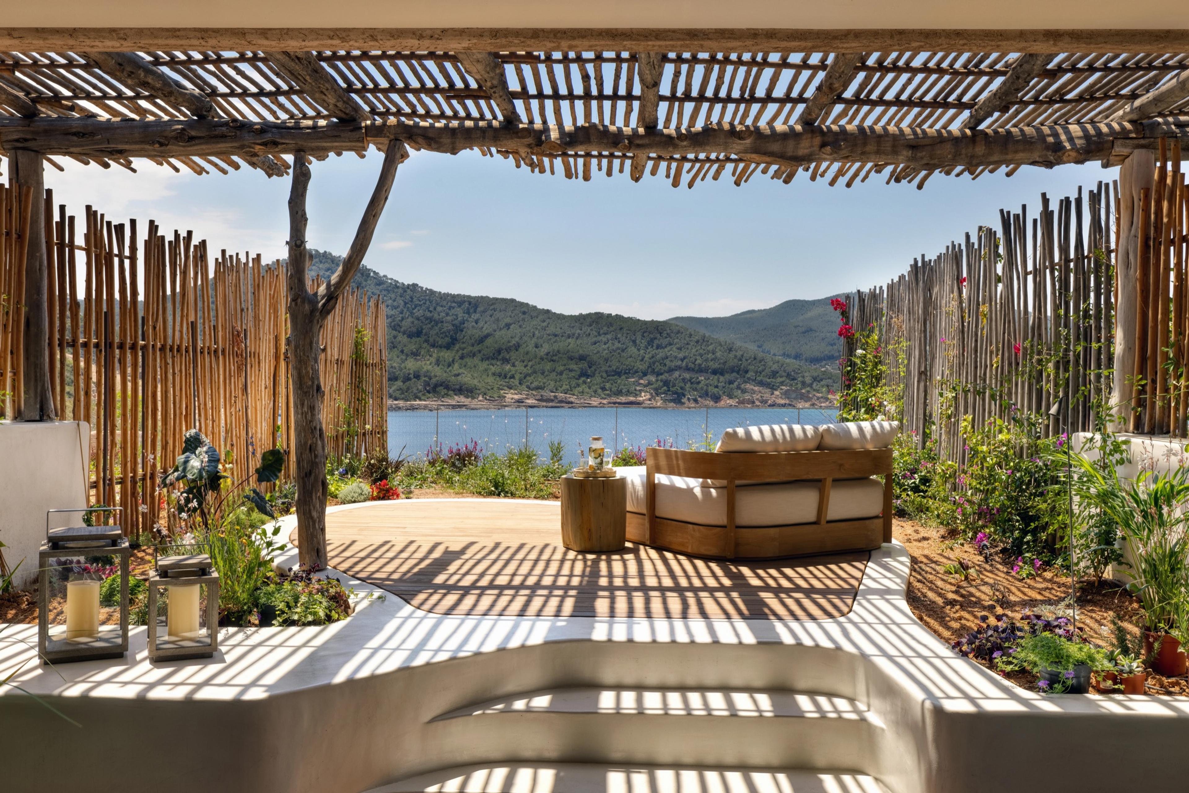 A raised outdoor lounge with a white sofa on a timber deck, enclosed by split-bamboo fencing and flowering plants, looking over a bay to wooded hills beyond.