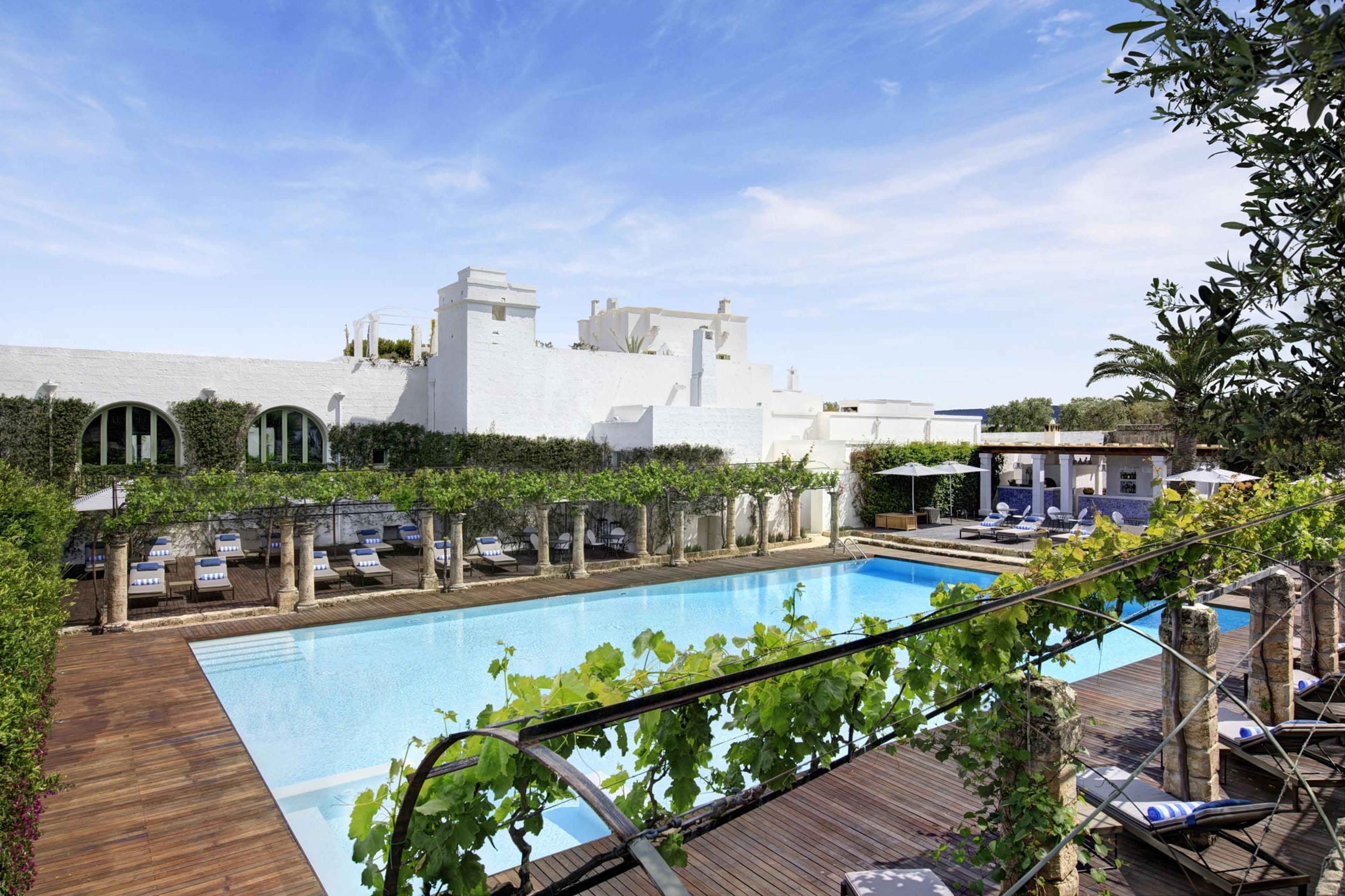 A rectangular swimming pool on a wooden deck beside a vine covered stone pergola, with a white building behind it under a blue sky.