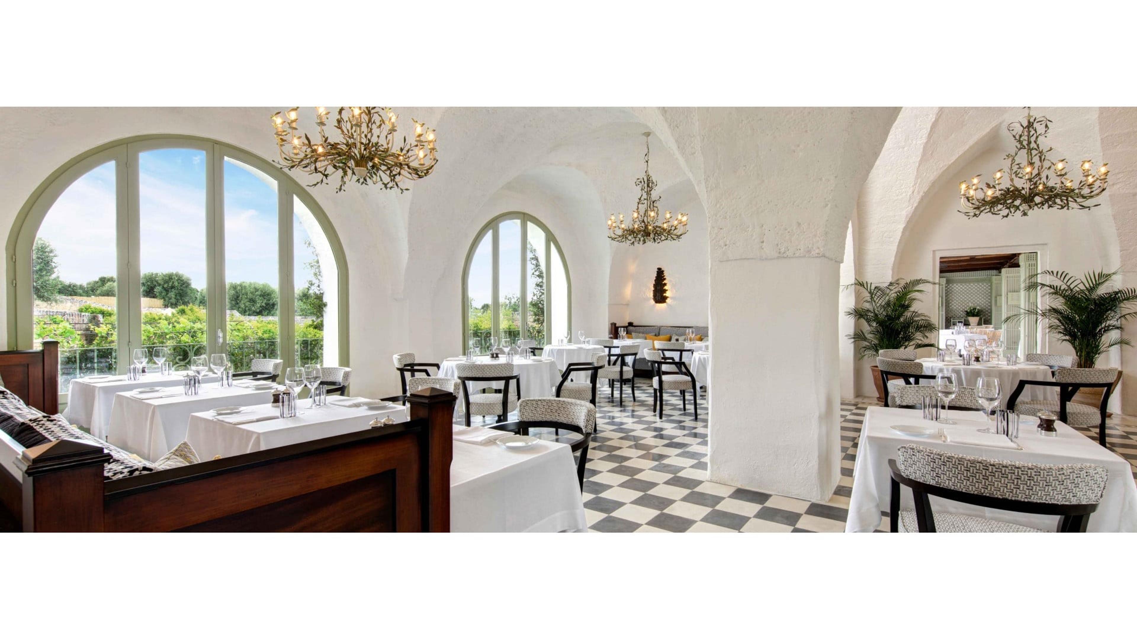 A vaulted white dining room with wrought iron chandeliers, arched windows, a black and white checkerboard floor and tables laid in white linen.