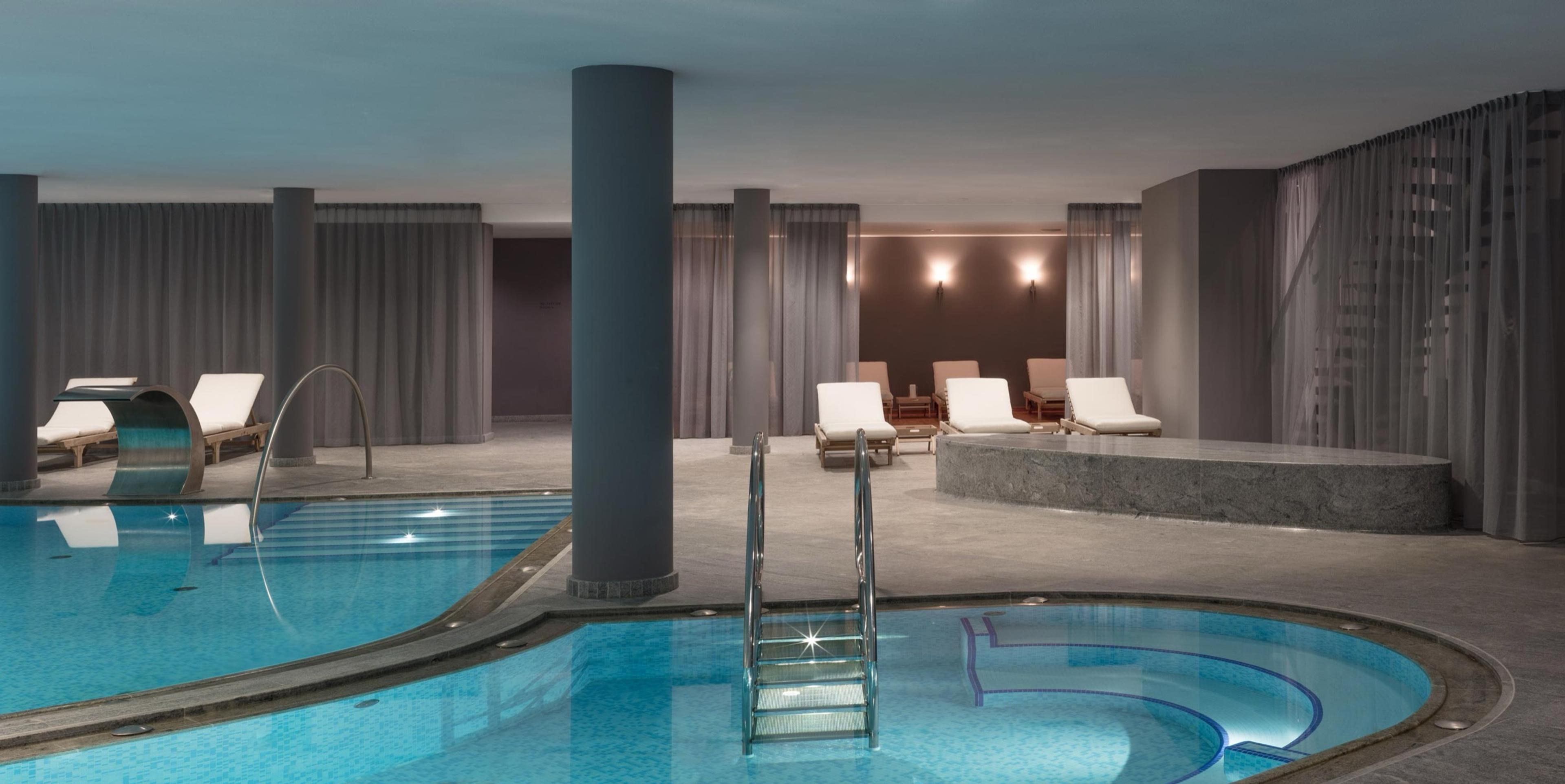An indoor hotel spa with a lap pool and a round plunge pool with steps, loungers and columns under dim lighting.