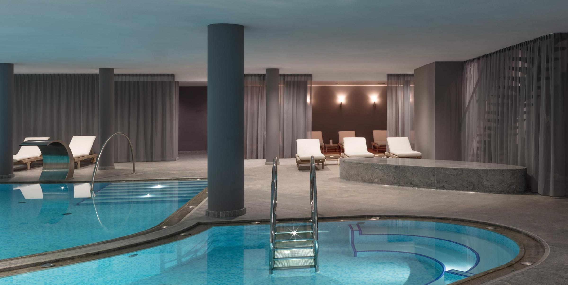 An indoor hotel spa with a lap pool and a round plunge pool with steps, loungers and columns under dim lighting.