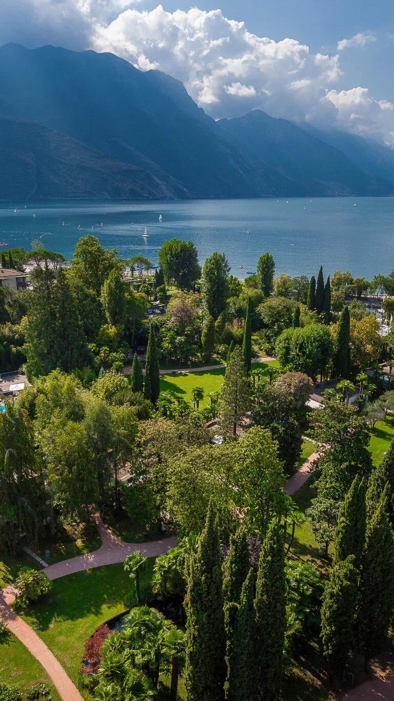 Aerial view down a garden of paths, lawns and tall cypresses, with a lake and mountains beyond.