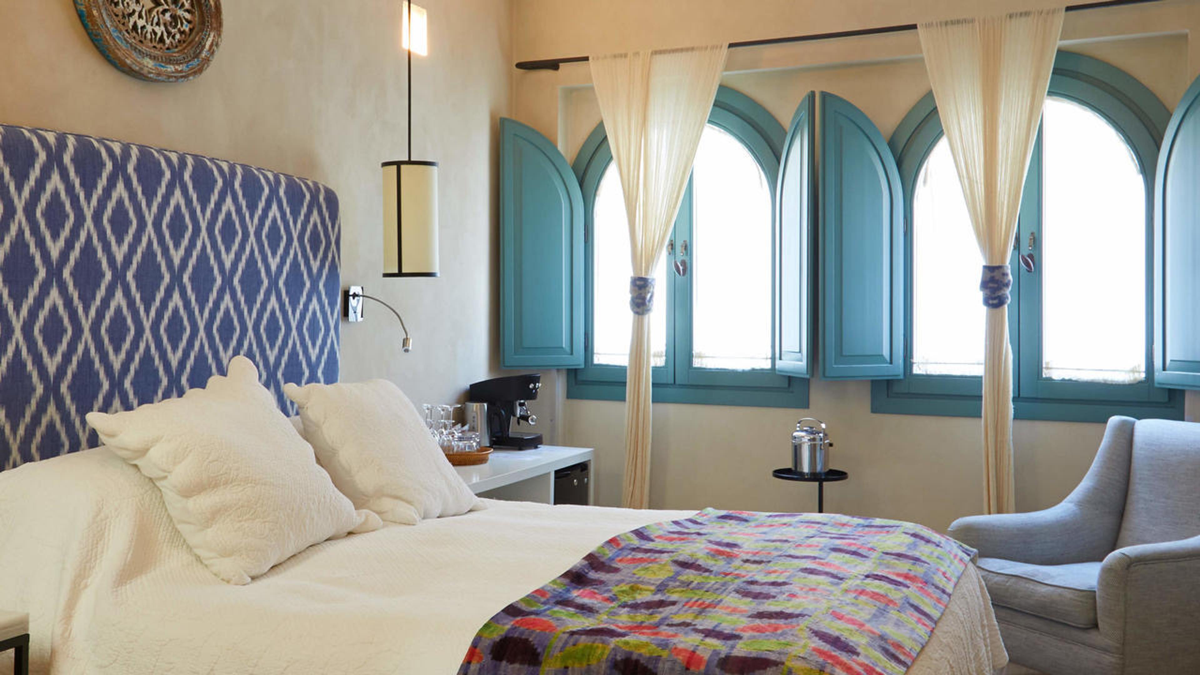 A bedroom with a blue diamond-patterned headboard, white bedding, a patterned throw, and a row of arched teal-shuttered windows with pale curtains.