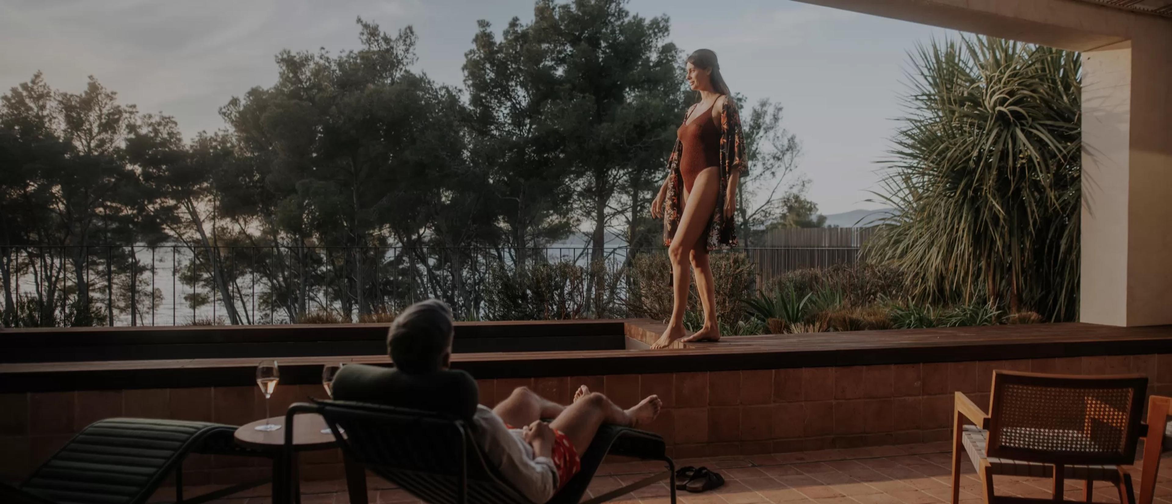 A woman in a red swimsuit and a sarong stands on a raised terrace under pine trees at dusk, while a man in a lounger in the foreground holds a glass of wine.