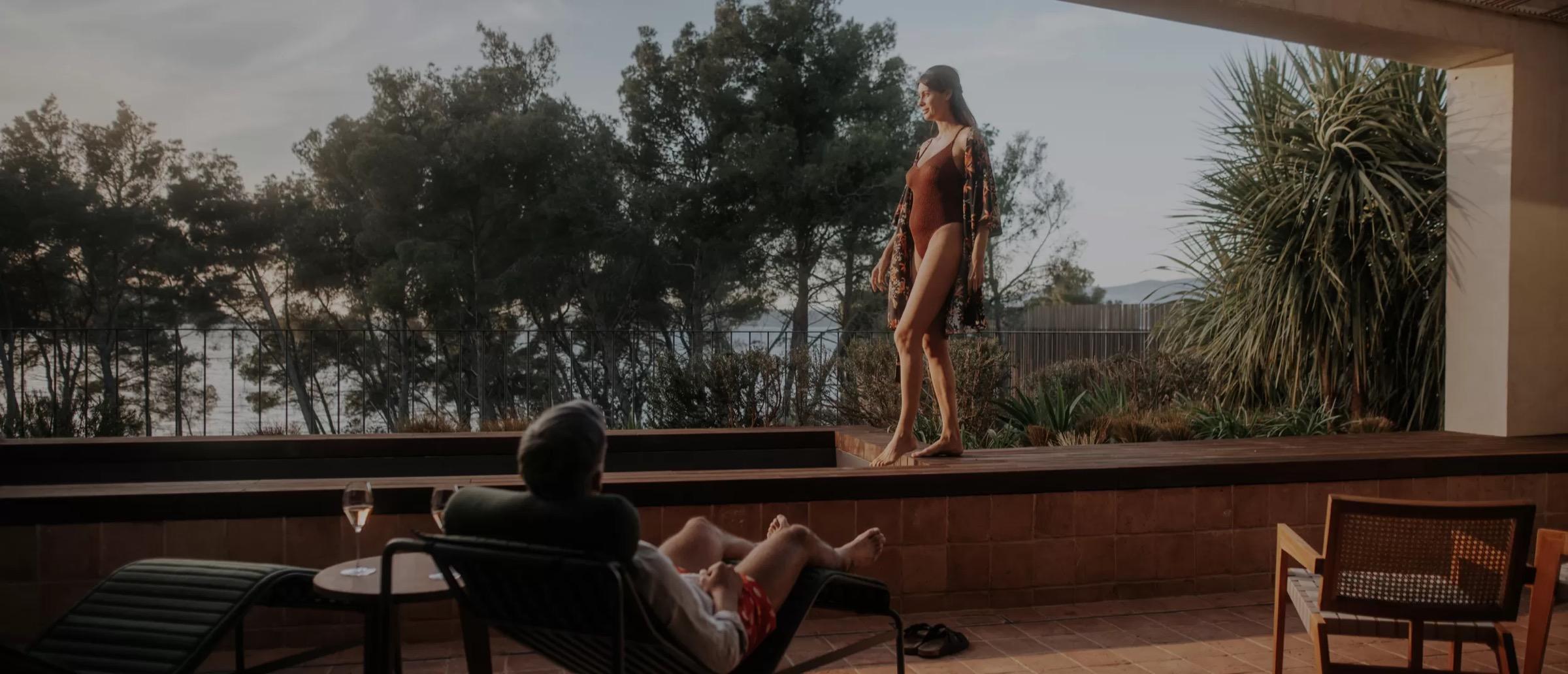 A woman in a red swimsuit and a sarong stands on a raised terrace under pine trees at dusk, while a man in a lounger in the foreground holds a glass of wine.