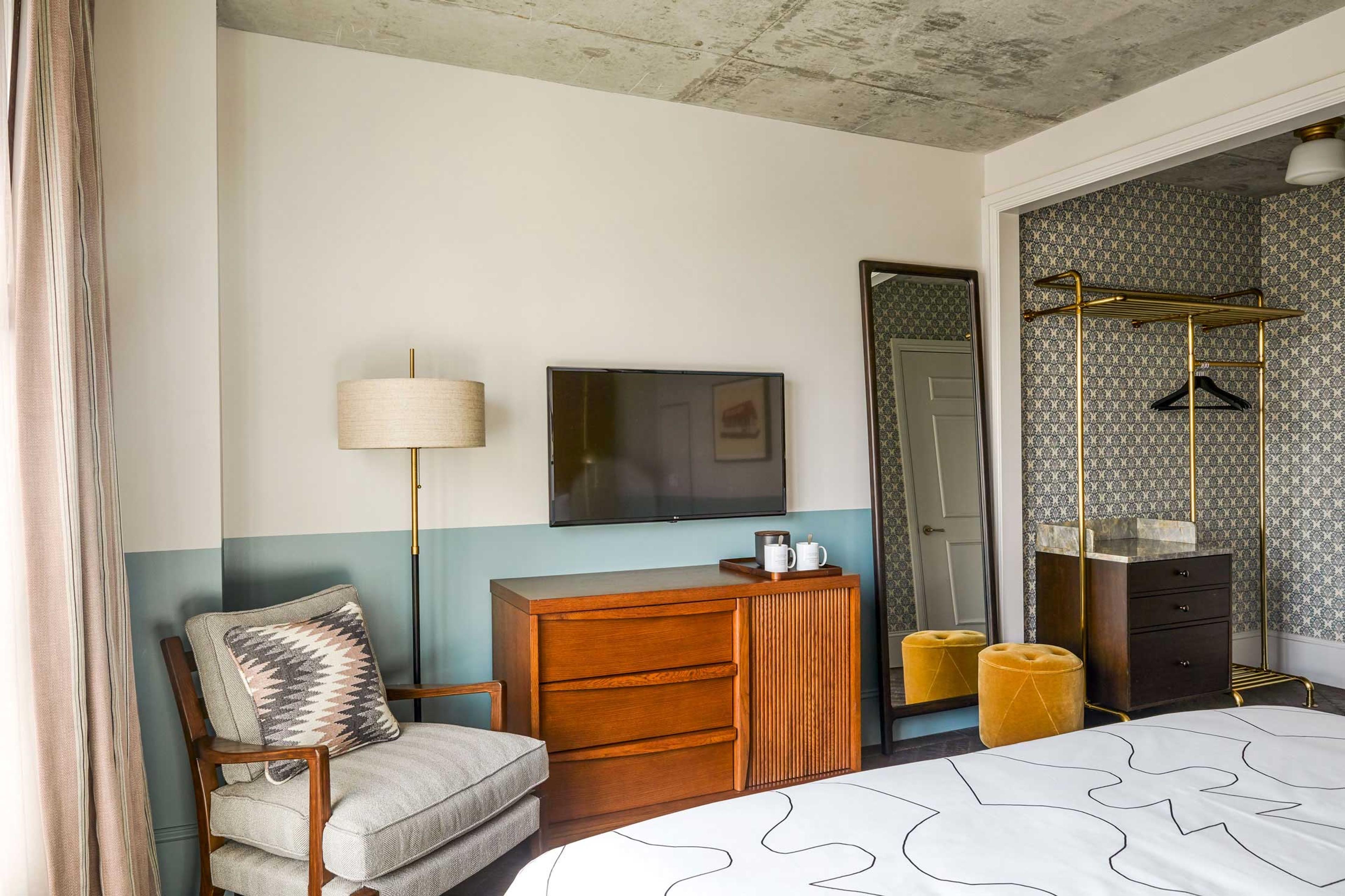 A hotel room with an exposed concrete ceiling, a wall mounted television above a wooden dresser, a tall mirror, an open clothes rail, and an armchair beside patterned curtains.