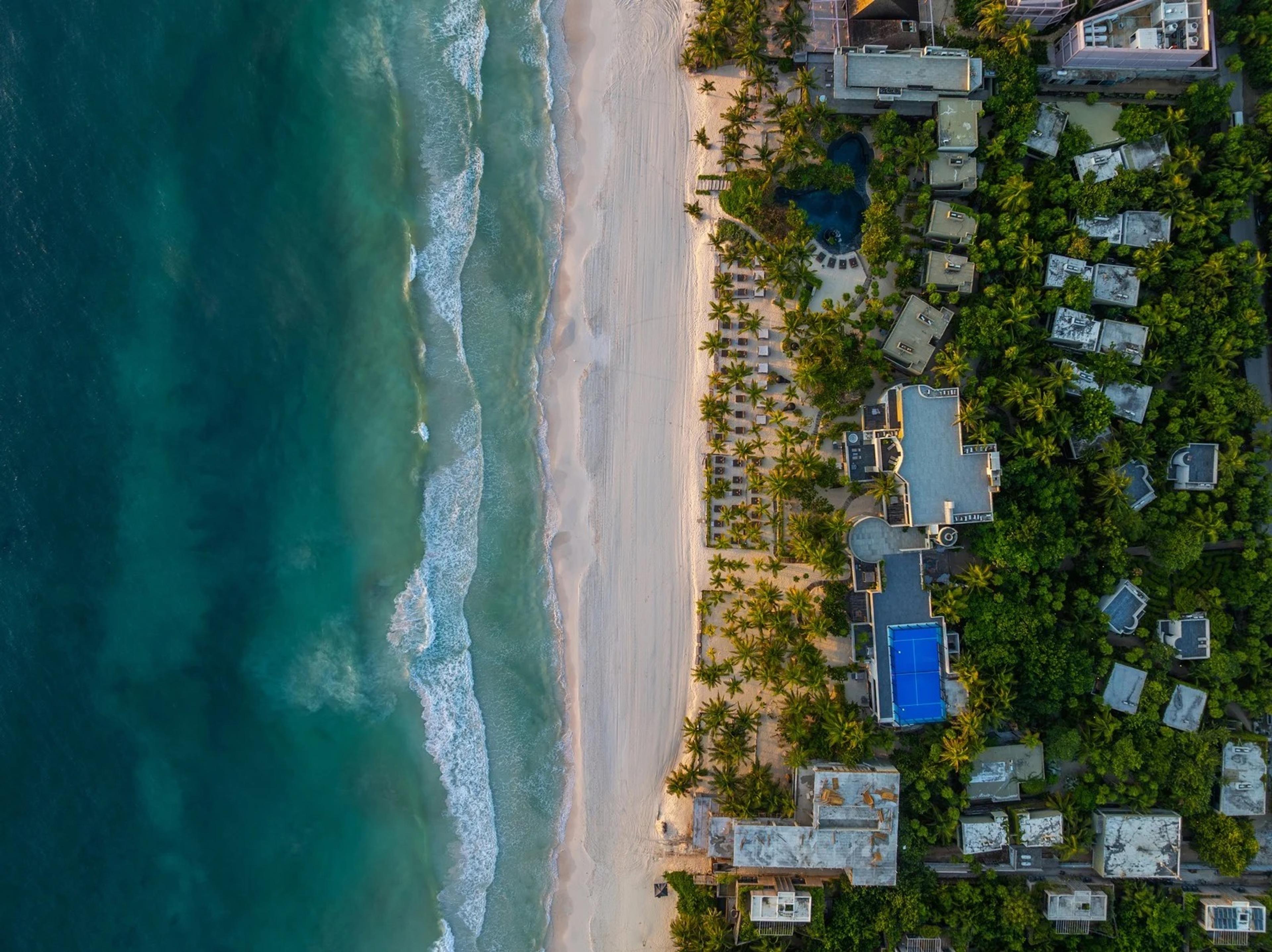 An aerial view of a beach resort, with turquoise sea, white sand, pools and palm-covered grounds.