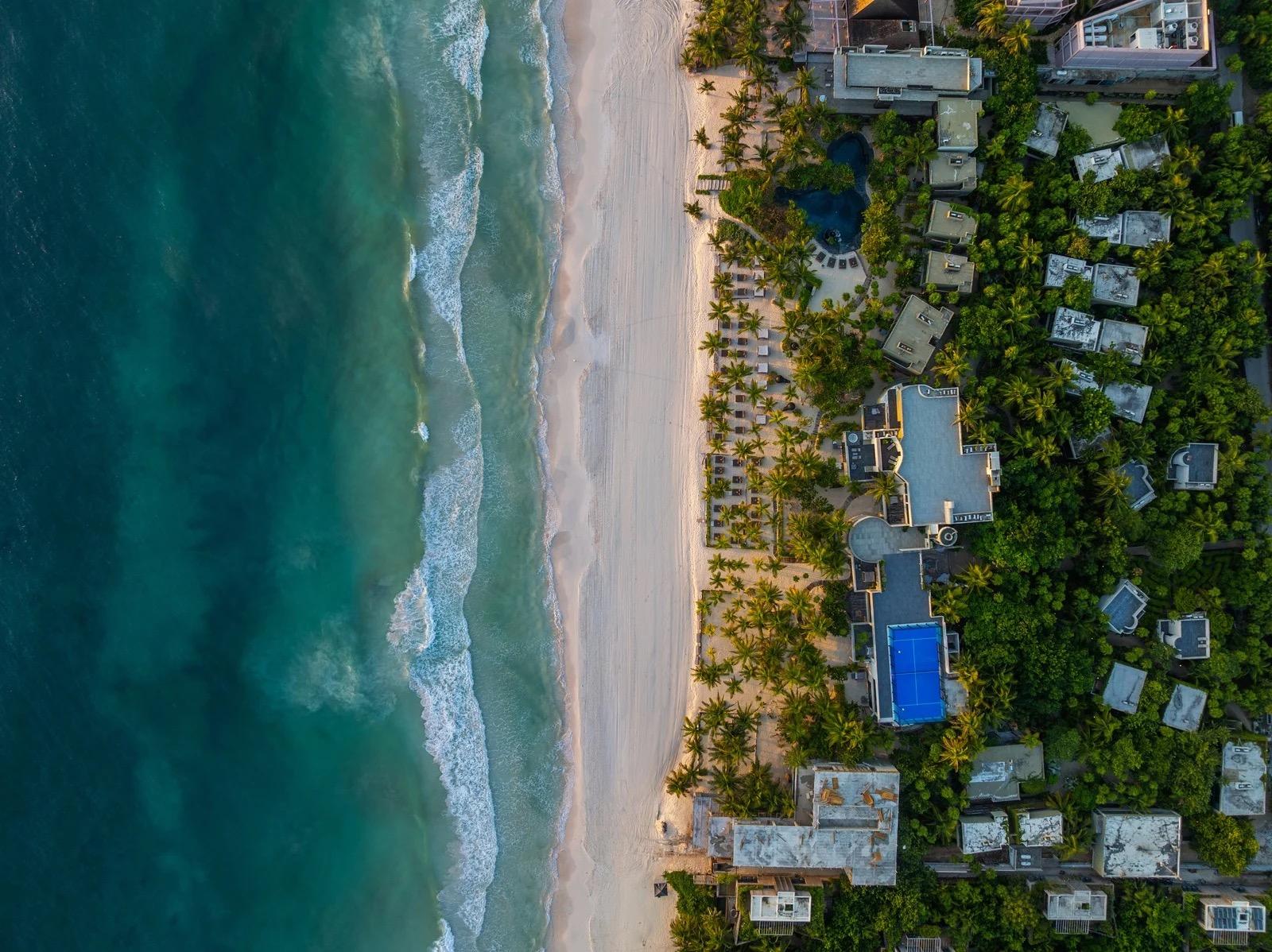 An aerial view of a beach resort, with turquoise sea, white sand, pools and palm-covered grounds.