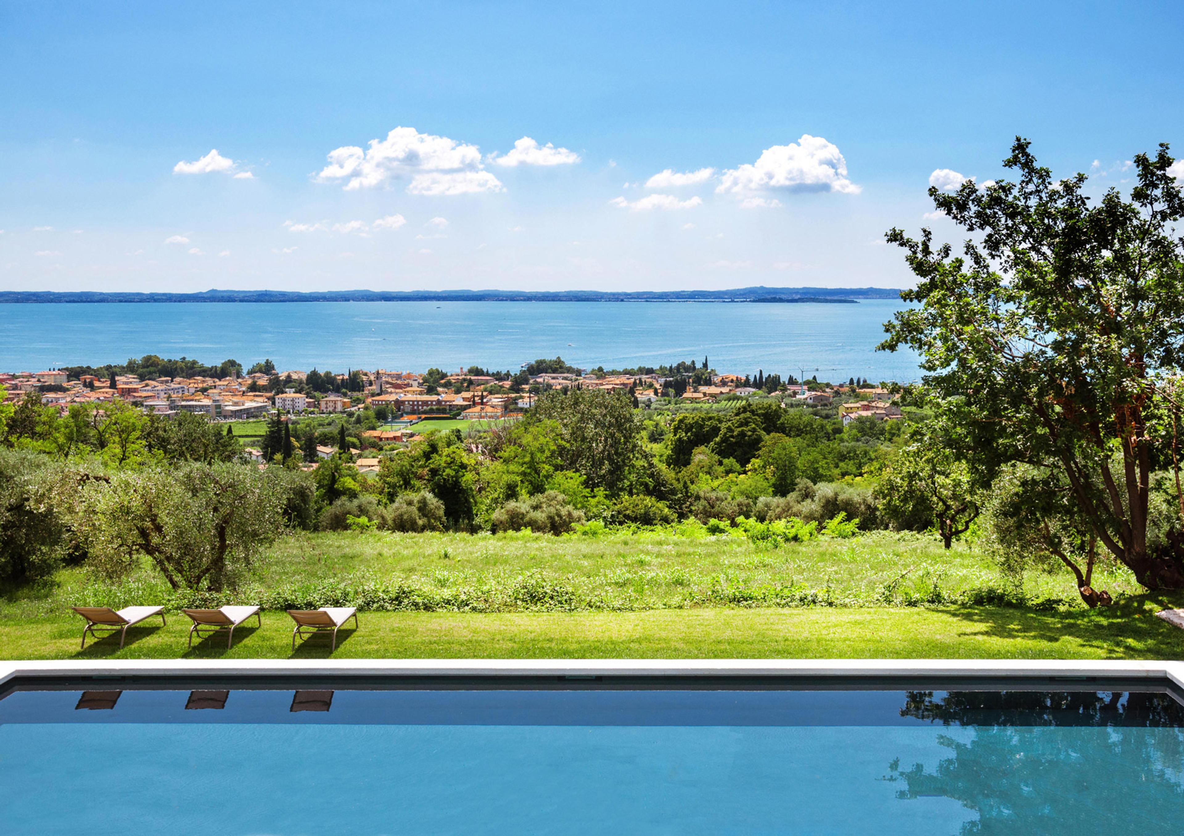 An infinity pool overlooking a lake and lakeside town, with three sun loungers on the grass beside it and trees to one side.