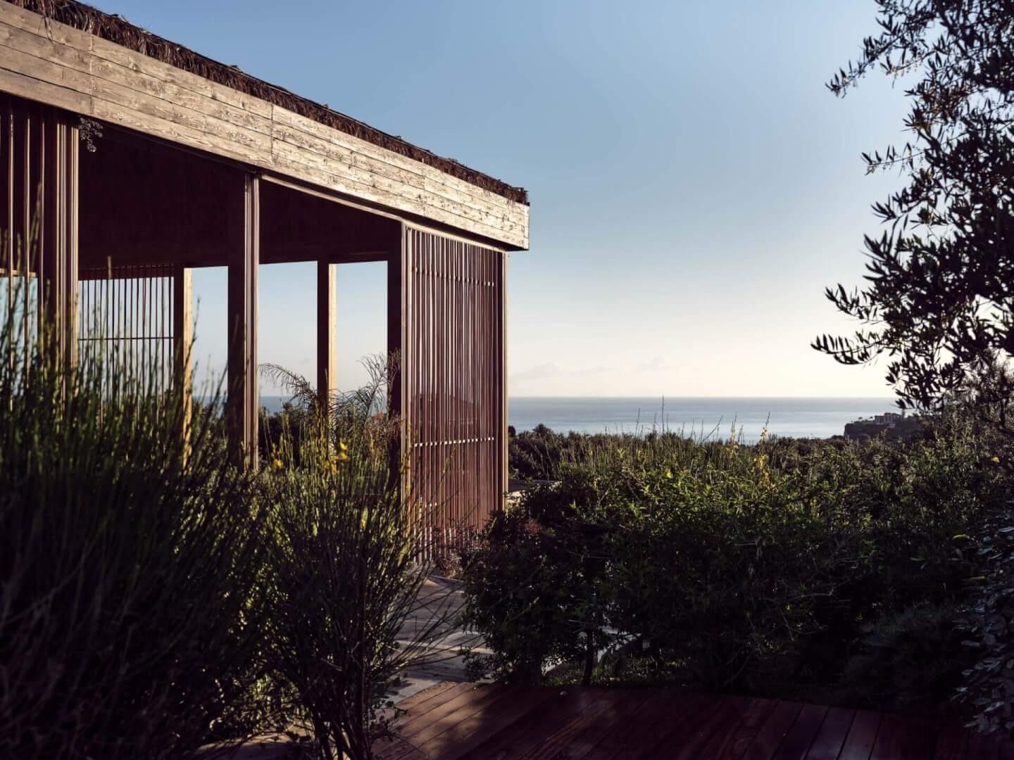 The timber-clad corner of a building with a slatted screen, framed by shrubs and olive branches with the sea visible beyond.