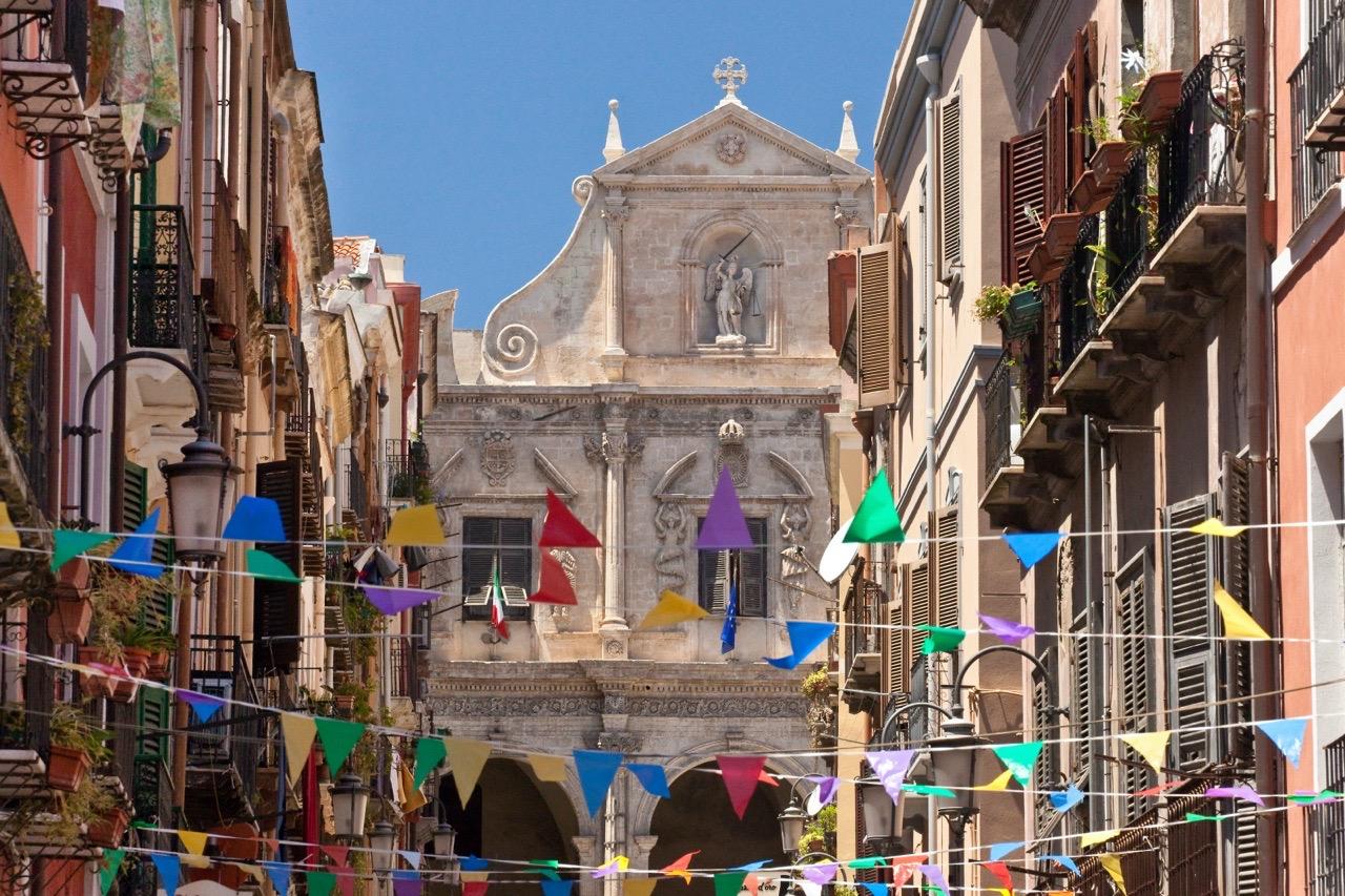 A narrow street strung with coloured triangular bunting, leading to a baroque church facade between balconied buildings, under a clear sky.