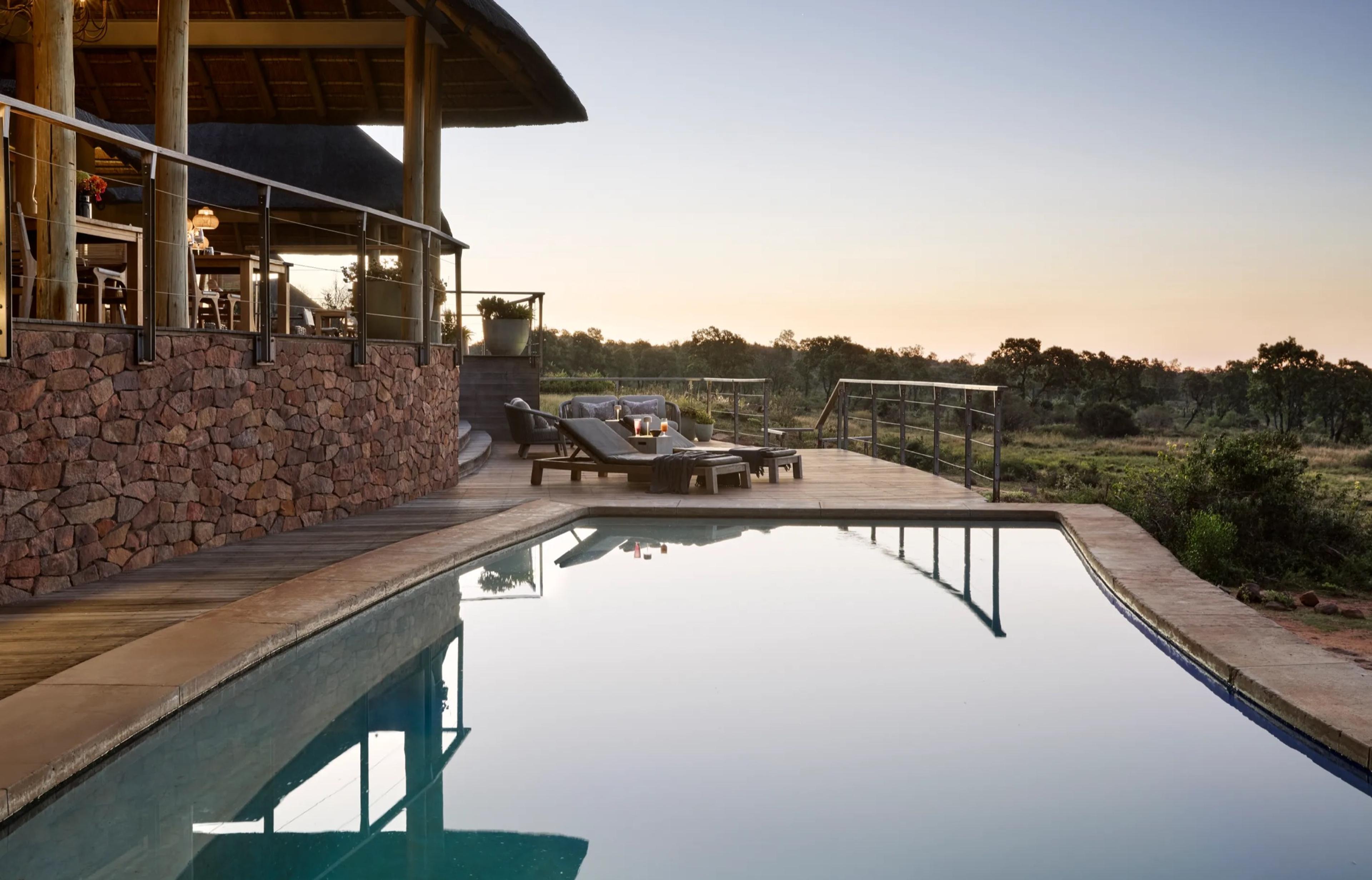 A pool at dusk beside a thatched lodge with a stone retaining wall, loungers on the deck, and bush and sky beyond.
