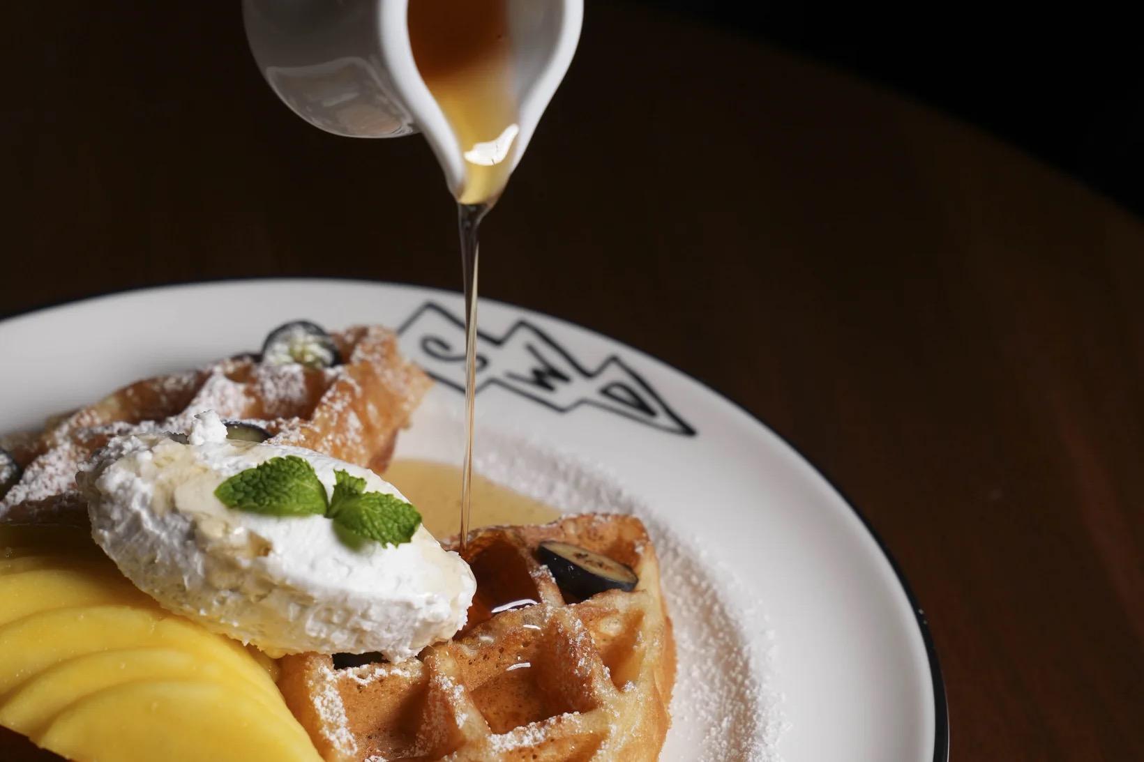 A waffle topped with whipped cream, mint, blueberries and mango slices on a plate, with syrup being poured over it from a small jug.