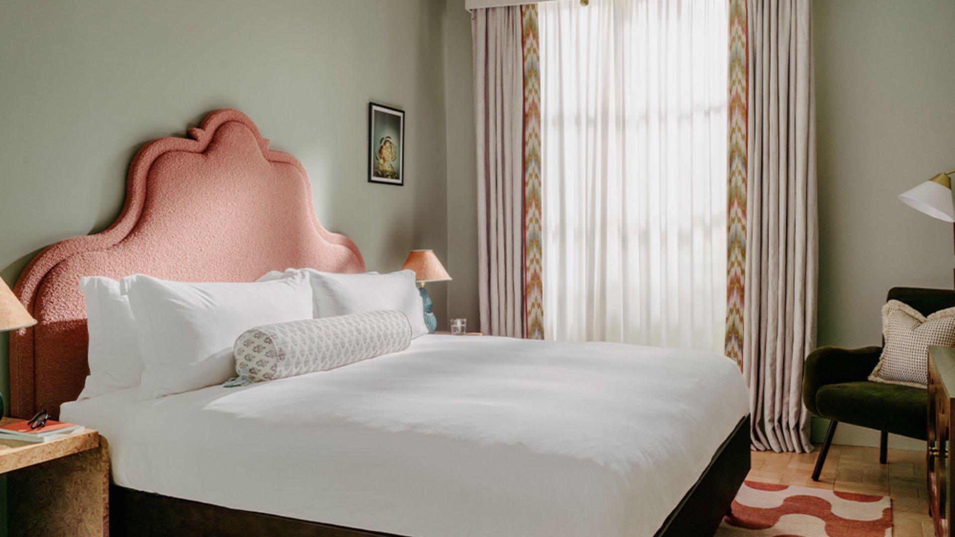 A bedroom with a pink scalloped headboard, patterned curtains at a tall window, a dark green armchair and a red and white patterned rug.