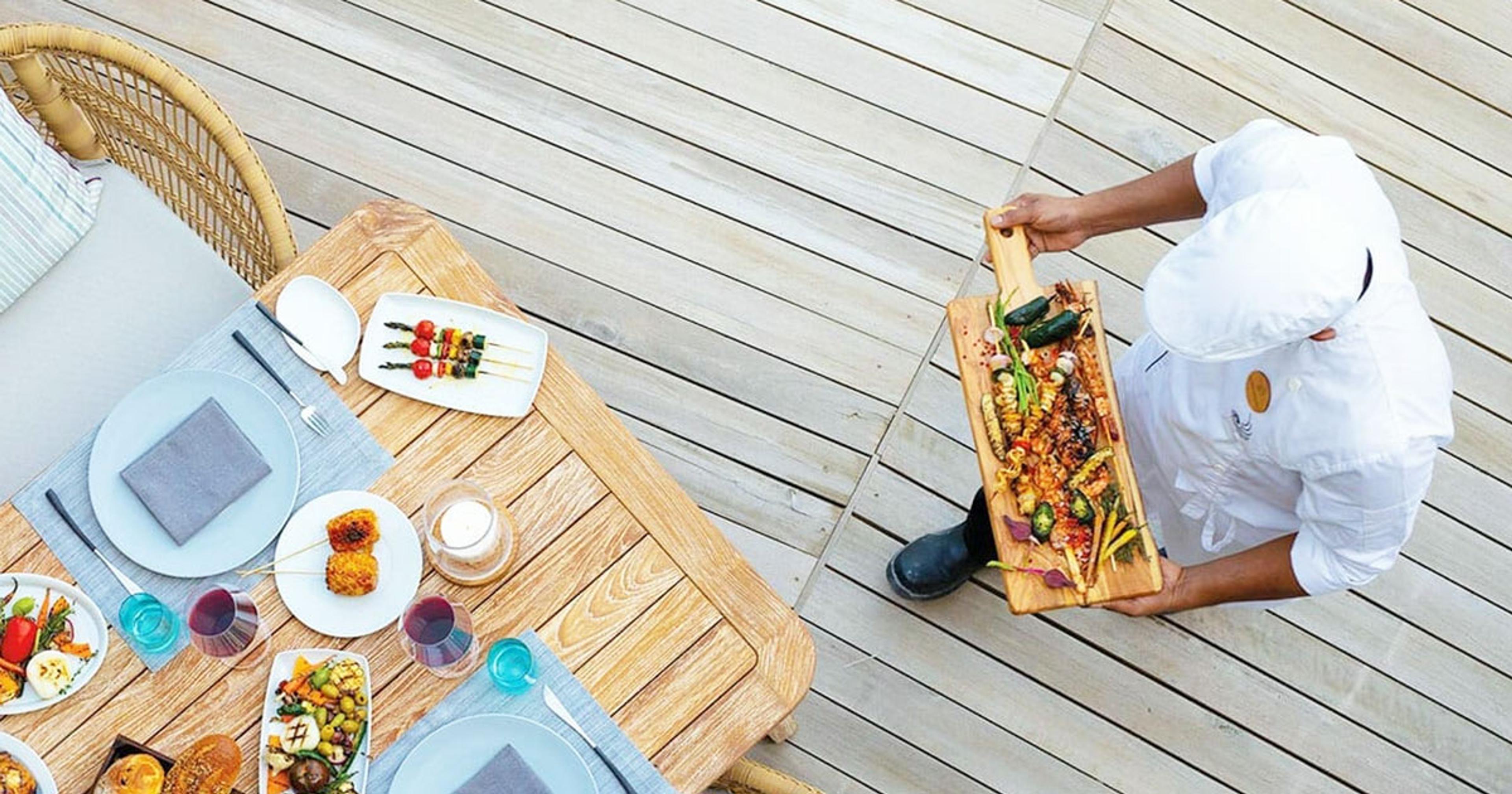 An overhead view of a chef setting down a board of grilled vegetables and meat beside a table laid with small plates and wine glasses.