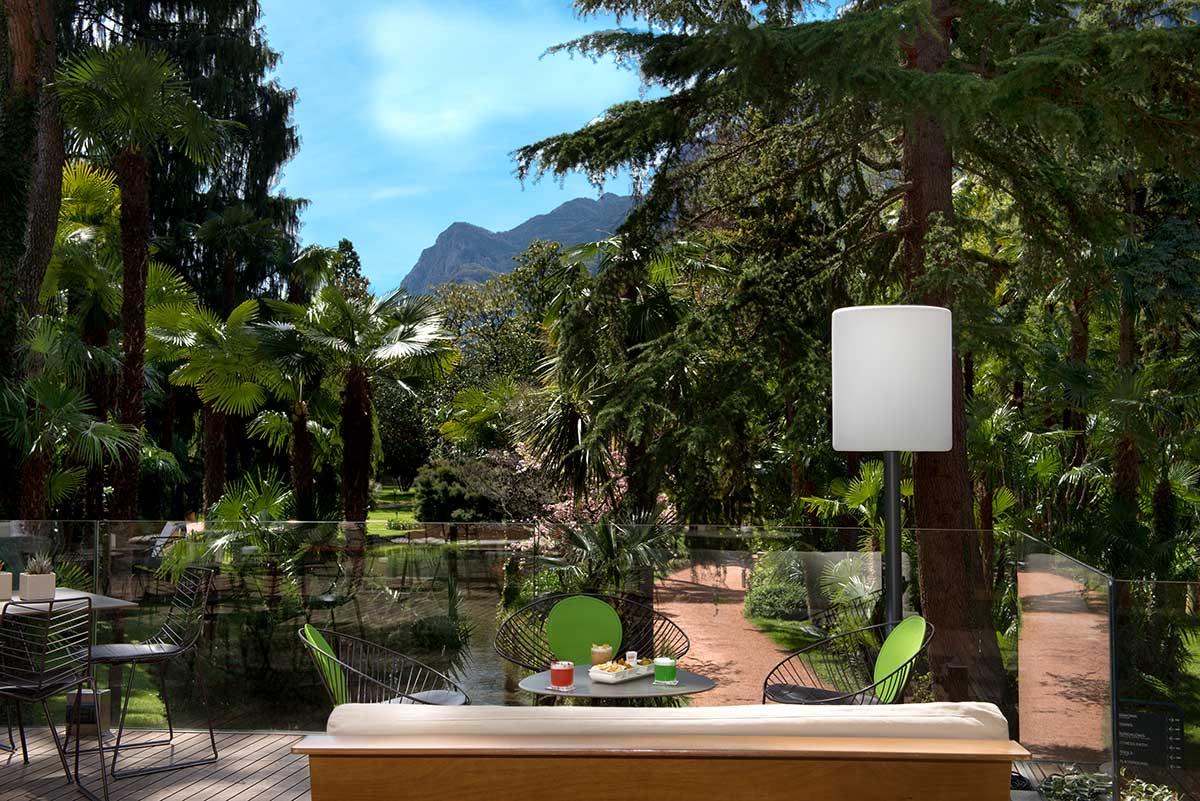 A glass-railed terrace with a cream sofa in the foreground, a white floor lamp, a small table with two drinks and a plate of snacks, and a view down through trees to a mountain.