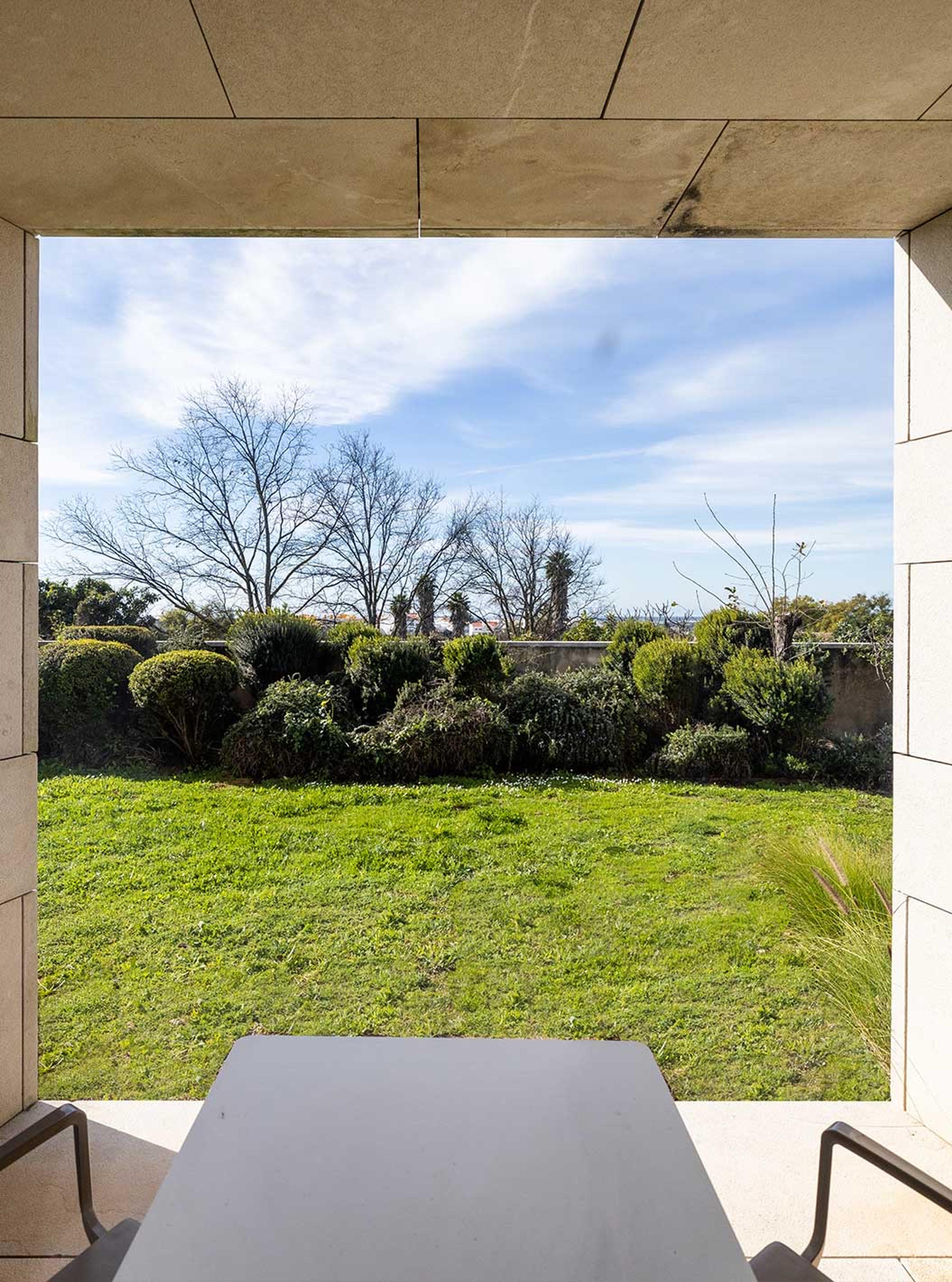 Through a stone-framed opening from a balcony, a grass lawn with shrubs and bare trees, and a white table in the foreground.