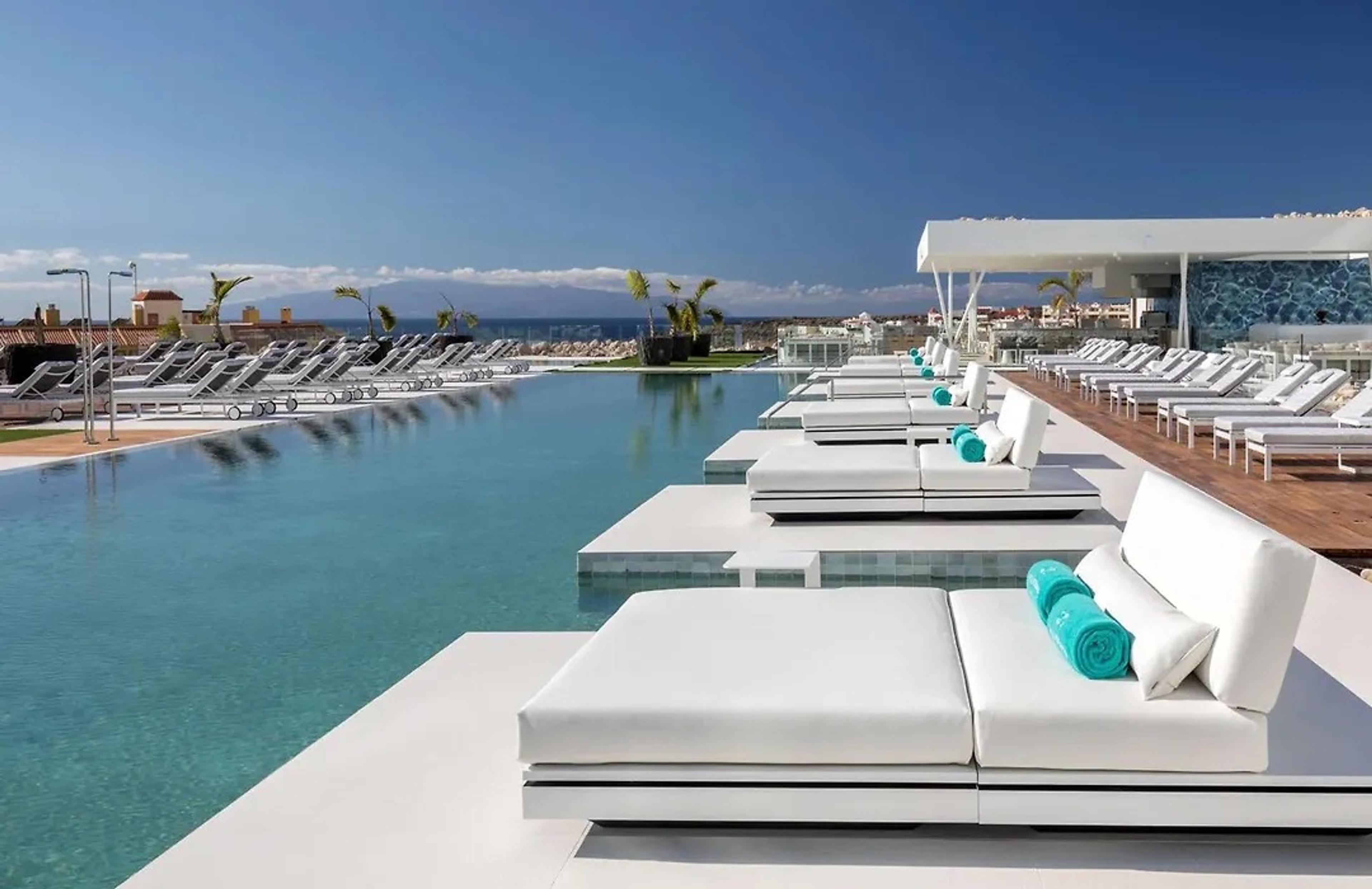 A long infinity pool lined with white daybeds and turquoise cushions, the sea and the coast of Tenerife beyond